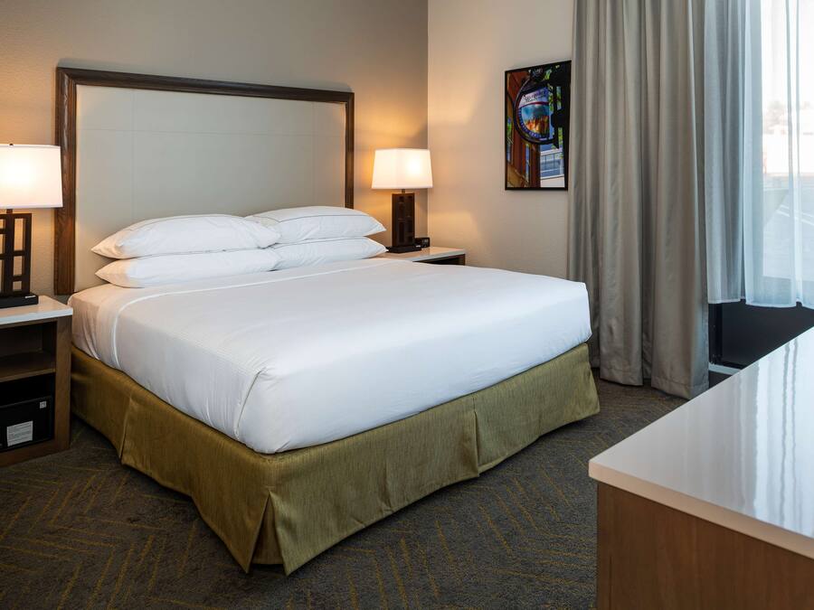 A hotel room with a large bed, two bedside tables with lamps, a carpeted floor, and a window with curtains.
