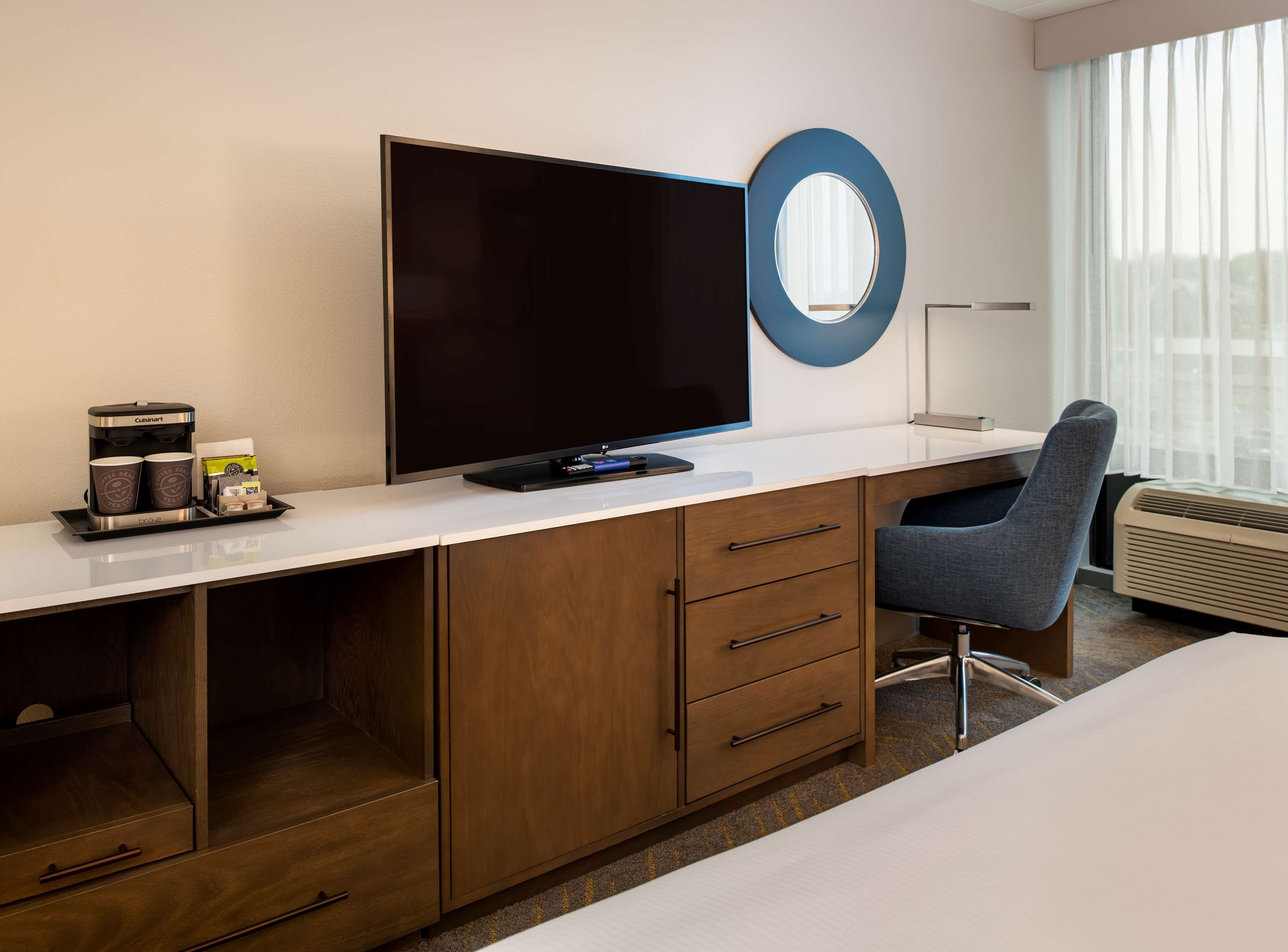 A hotel room with a large flat-screen TV, a coffee maker, a desk, a chair, and a large window with curtains.
