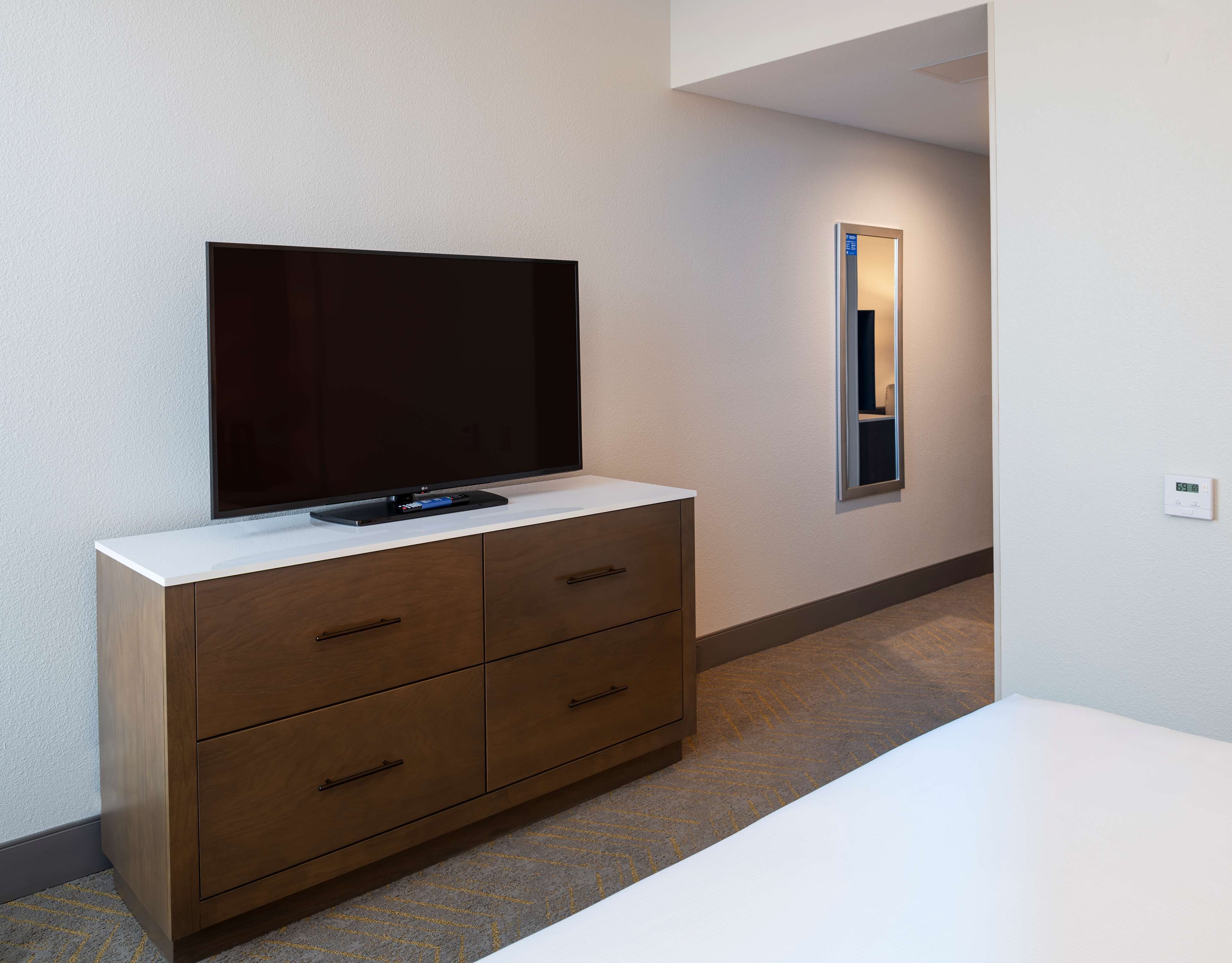A modern hotel room with a flat-screen TV on a wooden cabinet, a small mirror on the wall, and a thermostat on the wall.