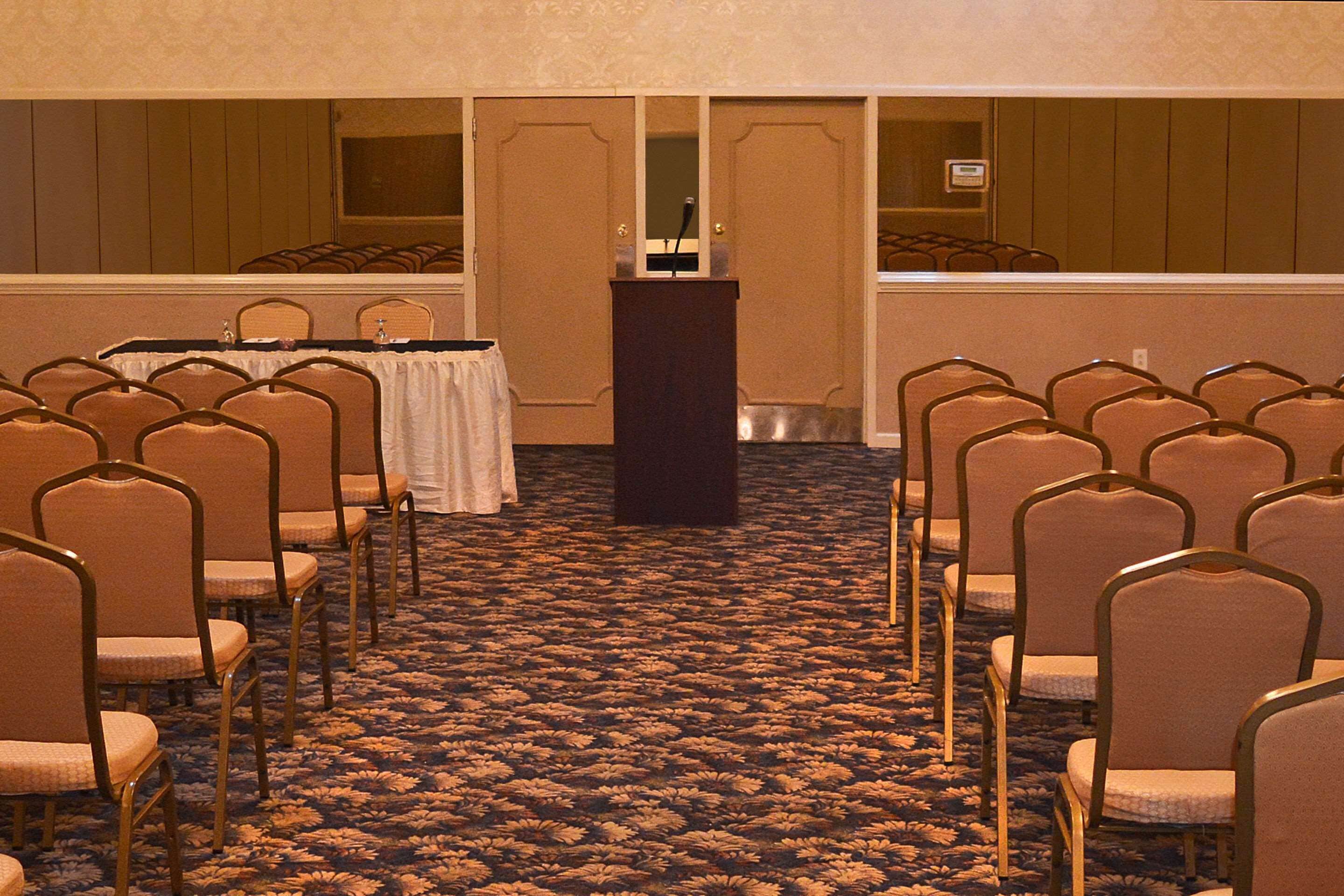 A conference room with rows of chairs facing a podium, carpeted floor, and wooden panel walls.