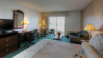 Quarto de hotel com uma cama grande, uma escrivaninha com computador, televisão, duas cadeiras, um sofá e uma janela com cortinas.