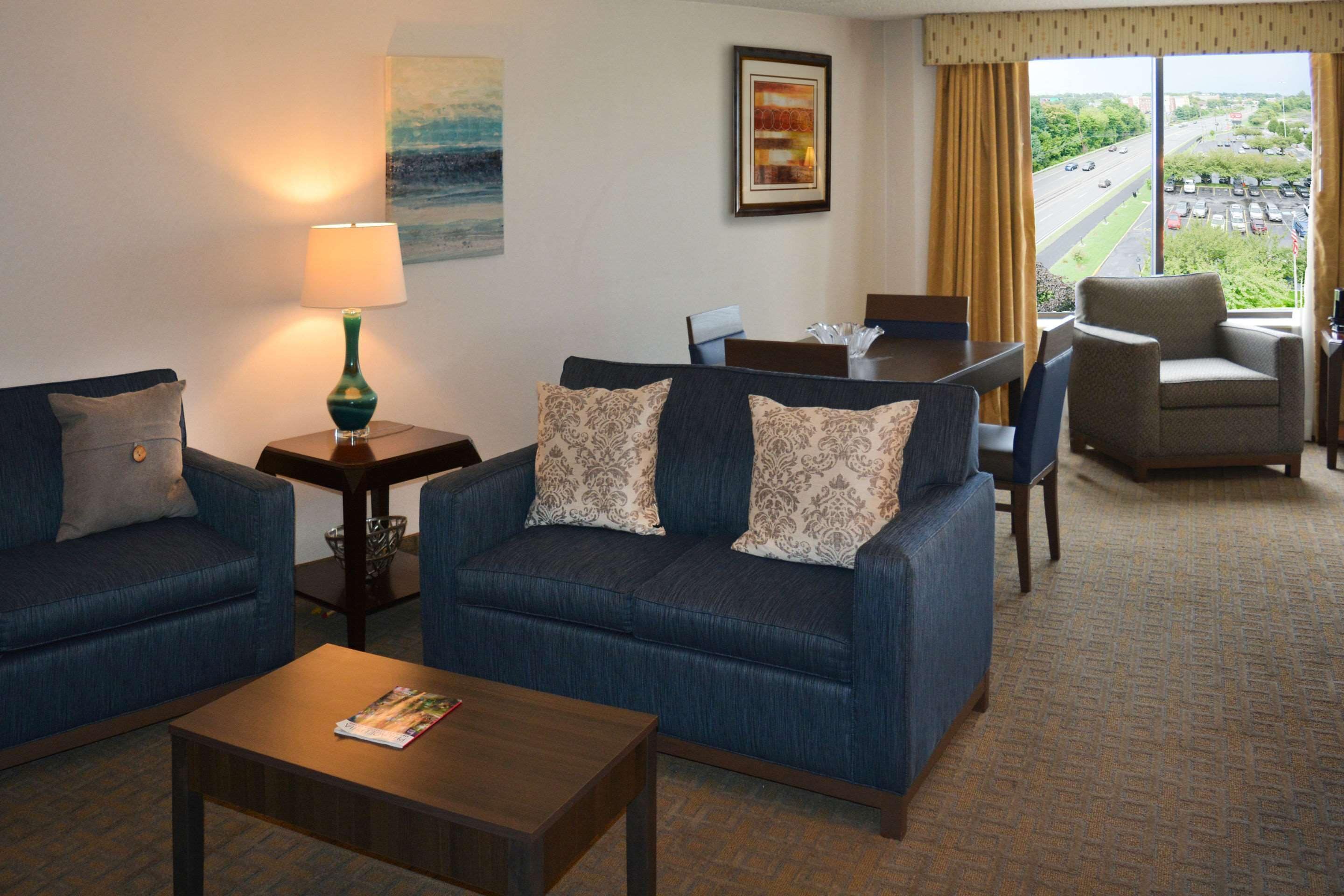 A hotel room with a blue sofa, a coffee table, a lamp, a dining table with chairs, and a view of a highway through large windows.