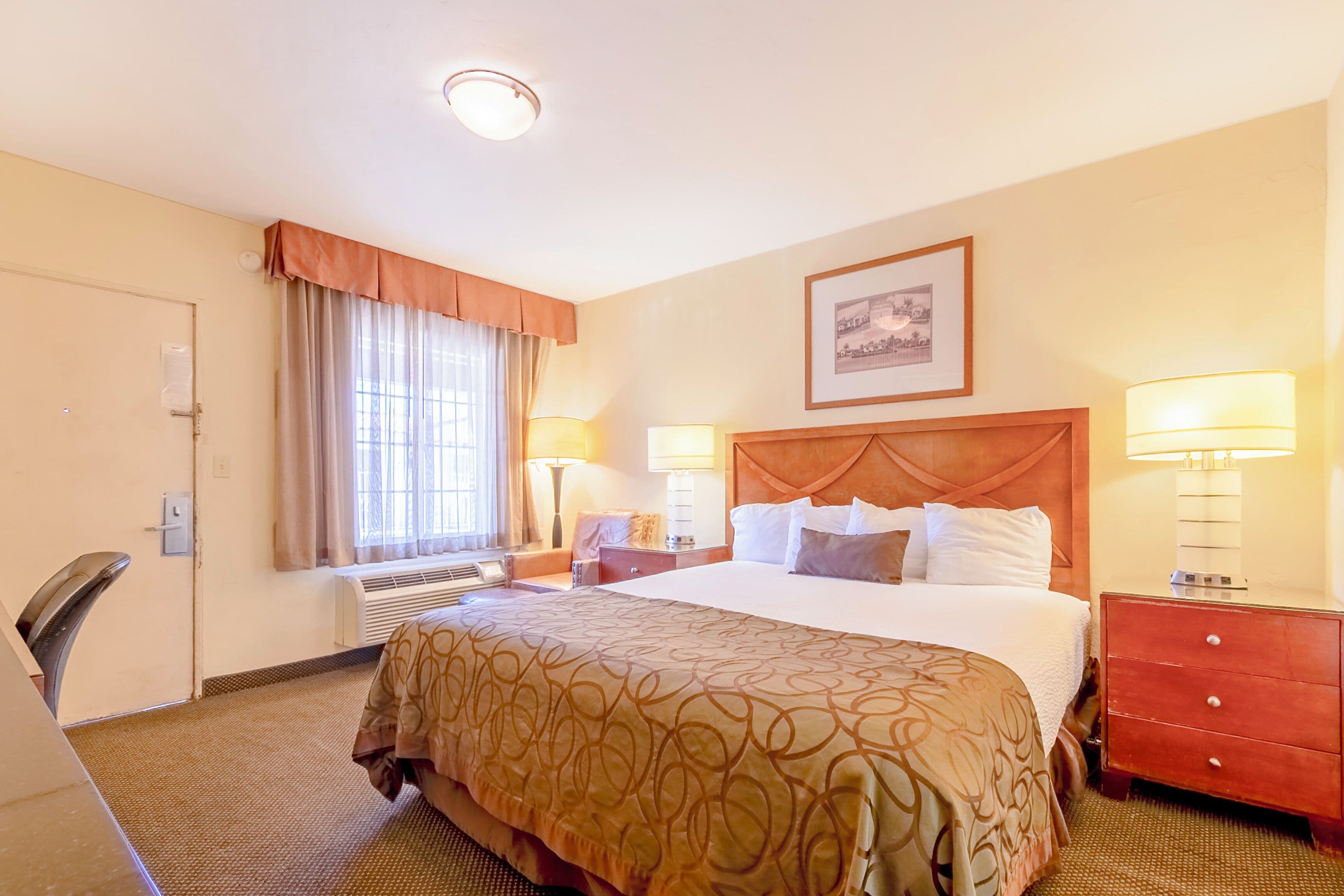 Une chambre d’hôtel avec un grand lit, deux tables de chevet avec des lampes, une tête de lit en bois, un tableau encadré au mur et une fenêtre avec des rideaux.