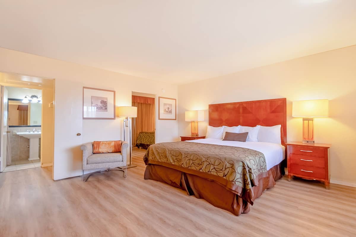 Una habitación de hotel con una cama grande, mesitas de noche con lámparas, una silla, un espejo y una puerta que da a un baño.