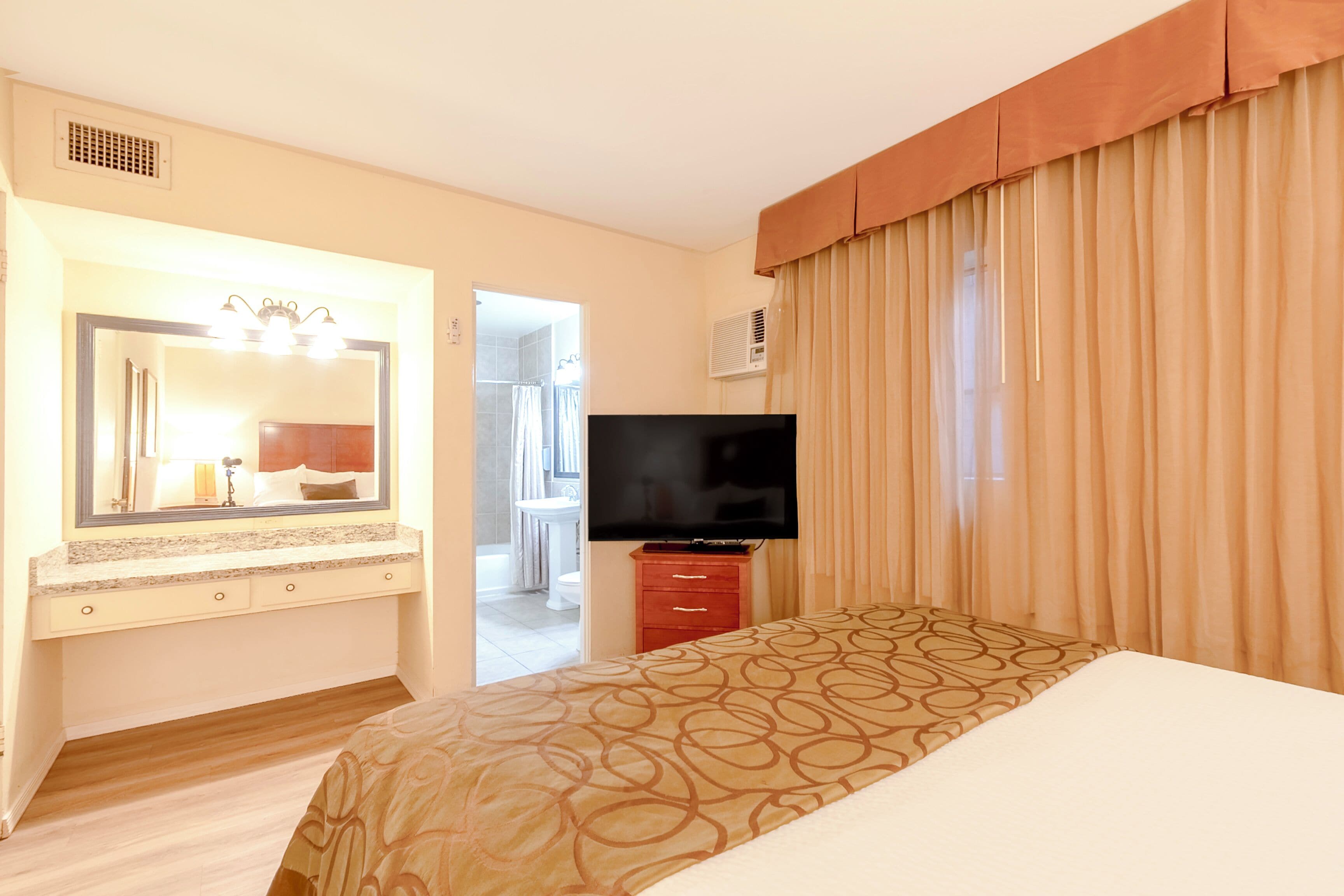 A hotel room with a bed, a TV, a vanity with a mirror, and a bathroom visible through an open door.