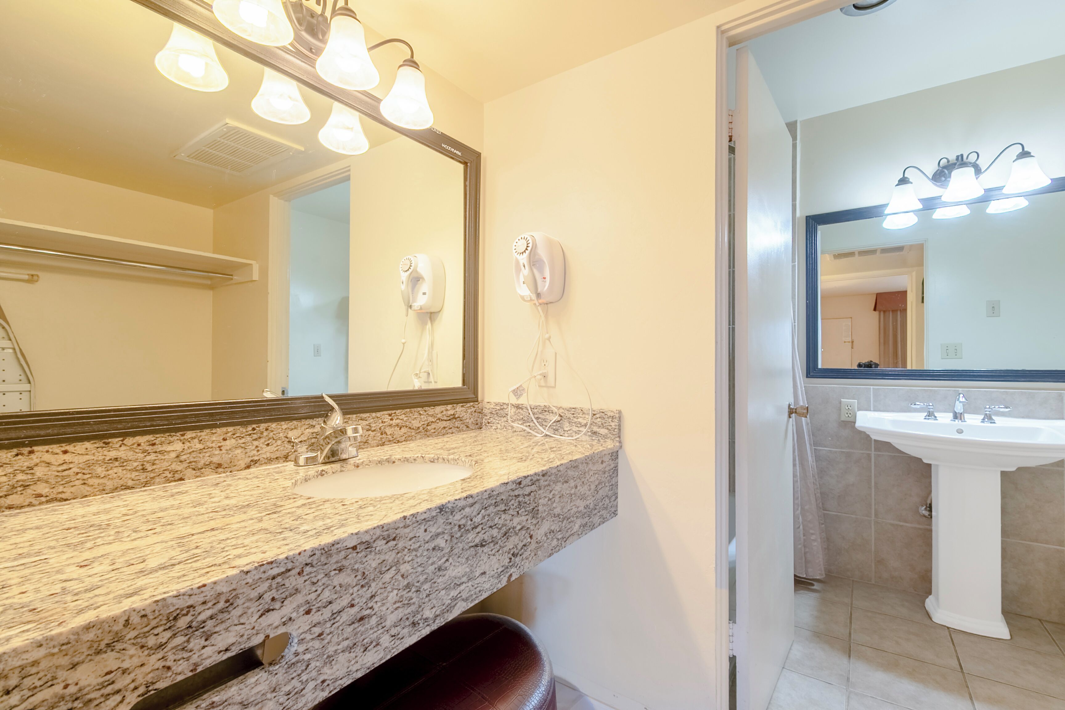 A bathroom with a granite countertop sink, a large mirror, and a pedestal sink. There are hair dryers mounted on the wall and a shower visible in the background.