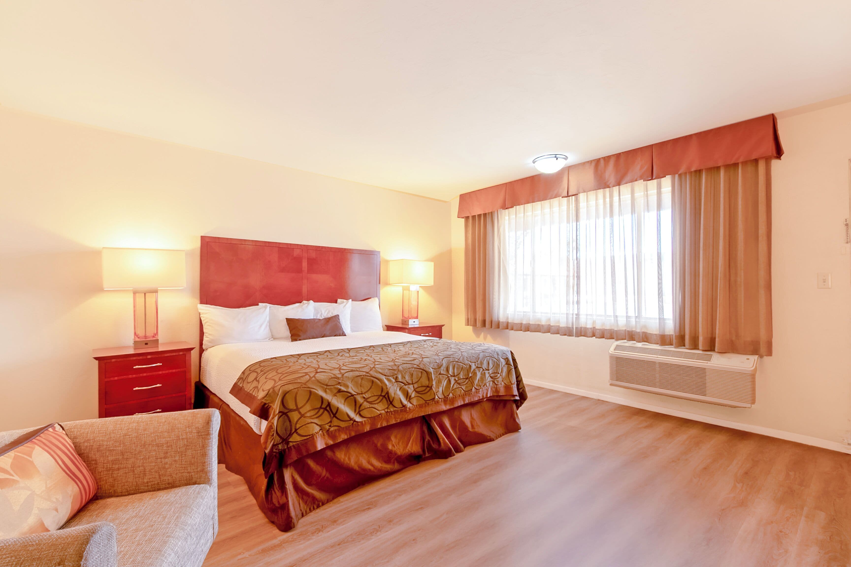A hotel room with a bed, bedside tables, a chair, a window with curtains, and an air conditioning unit.