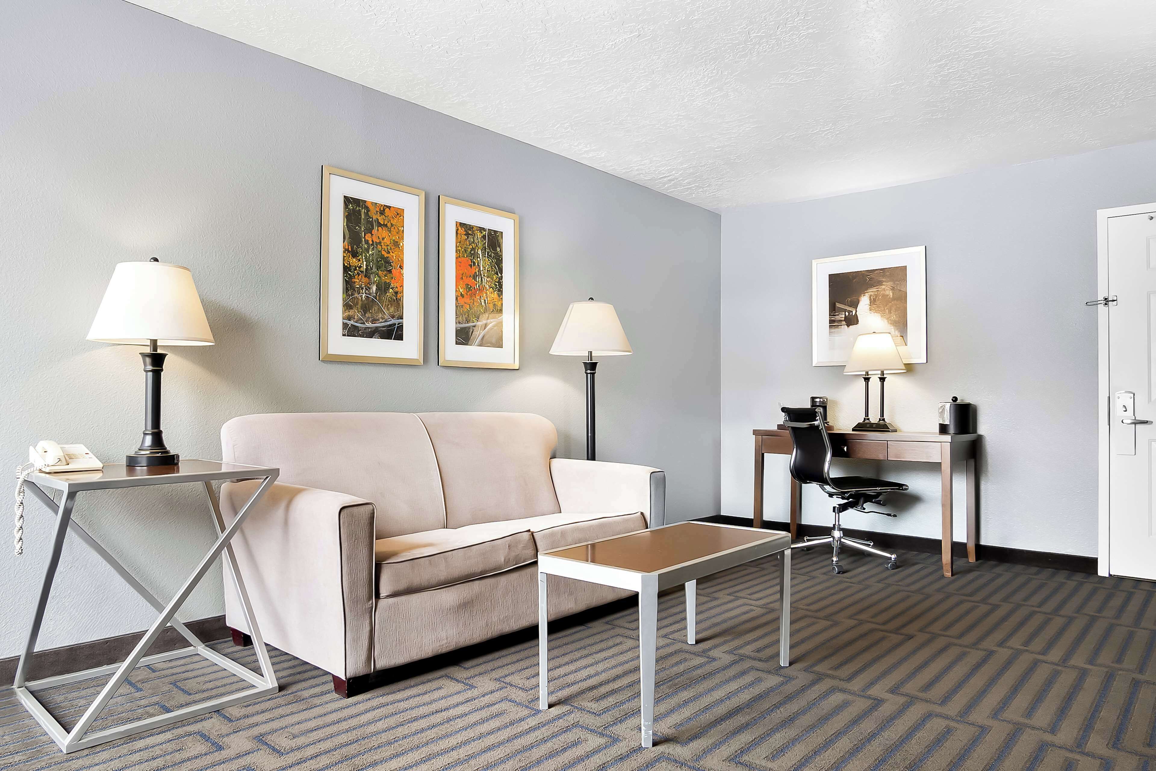 A hotel room with a sofa, a coffee table, two lamps, a desk, and framed pictures on the wall.