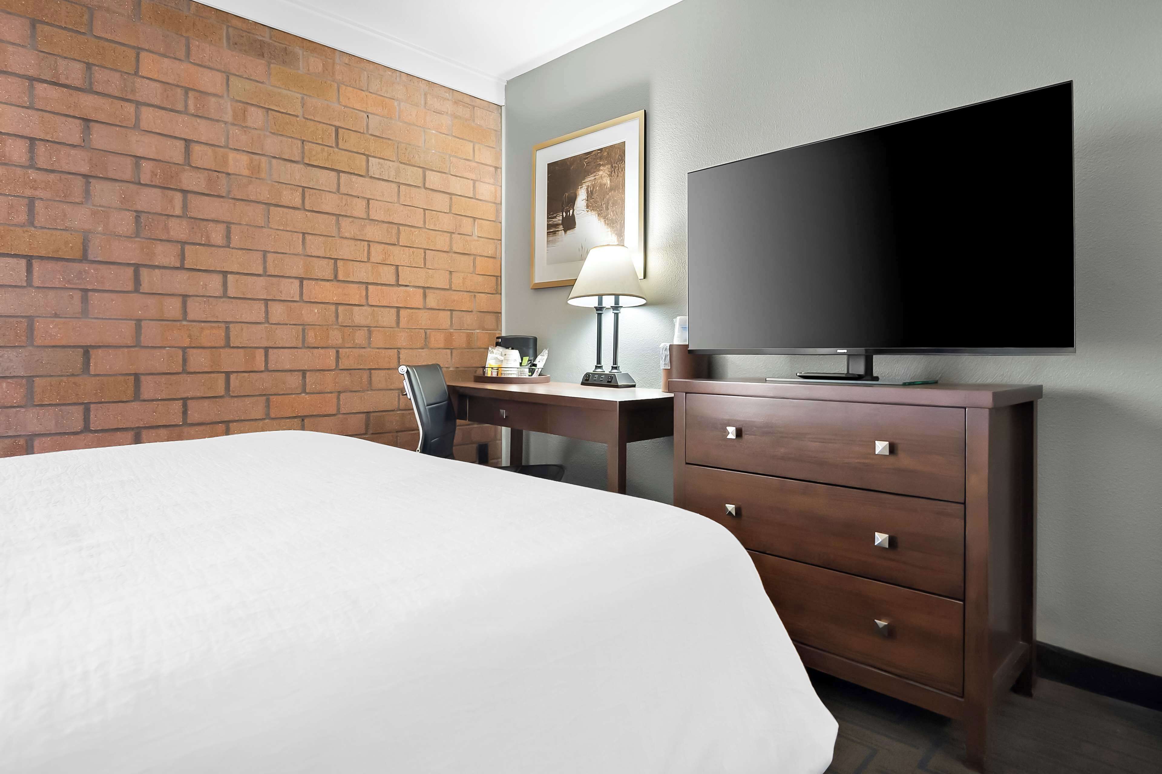 A hotel room with a brick wall, a bed, a desk with a lamp, a television, and a dresser.