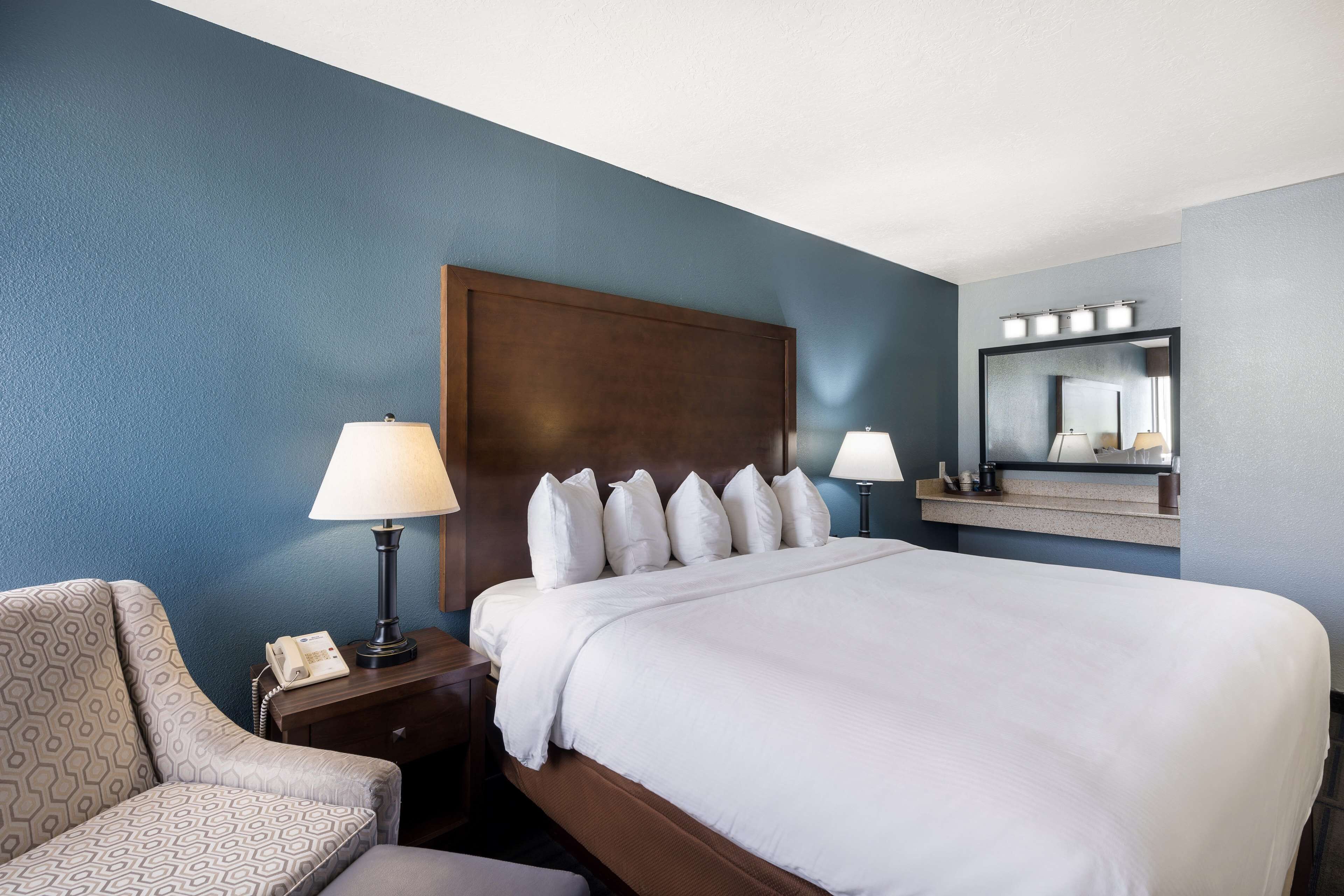 A hotel room with a large bed, two bedside lamps, a nightstand with a telephone, a chair, and a mirror above the bed.