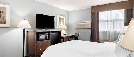 A hotel room with a bed, desk, chair, television, and air conditioning unit.