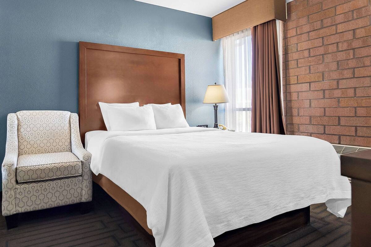 A hotel room with a large bed, a wooden headboard, a patterned armchair, a bedside table with a lamp, and a brick wall.