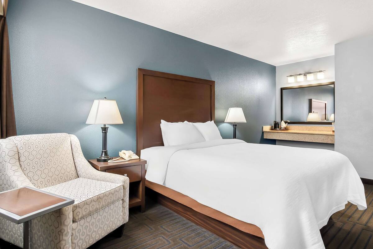 A hotel room with a bed, a chair, a nightstand with a lamp, and a mirror with lights.