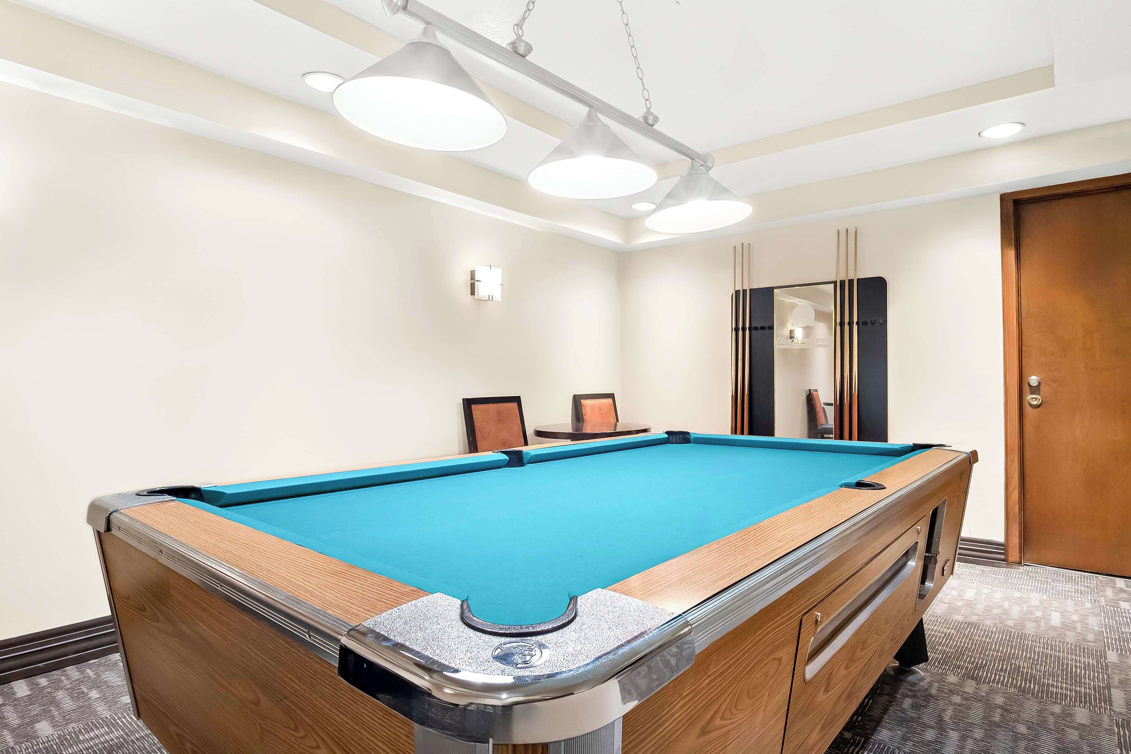 A pool table with a blue felt surface, a wooden frame, and a metal rail. There are overhead lights and a door in the background.