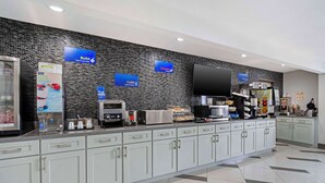 A modern hotel lobby with a self-service breakfast area, including a coffee machine, toaster, and refrigerated display.