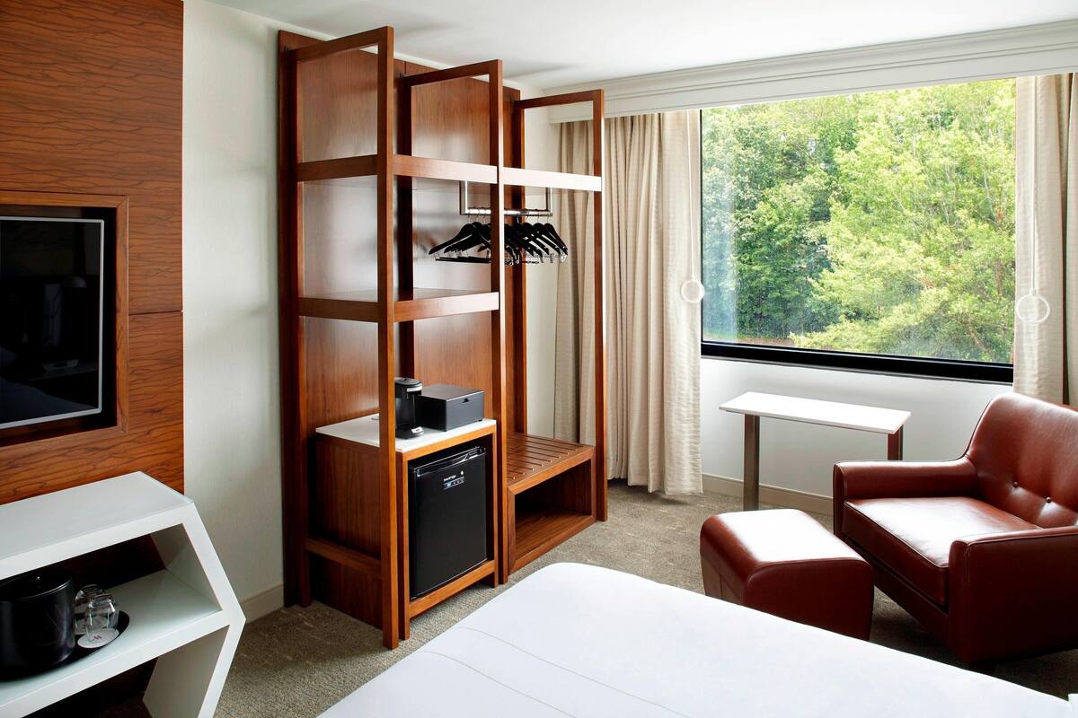 A hotel room with a bed, a wooden wardrobe, a small desk with a television, a red leather chair, and a window with a view of trees.