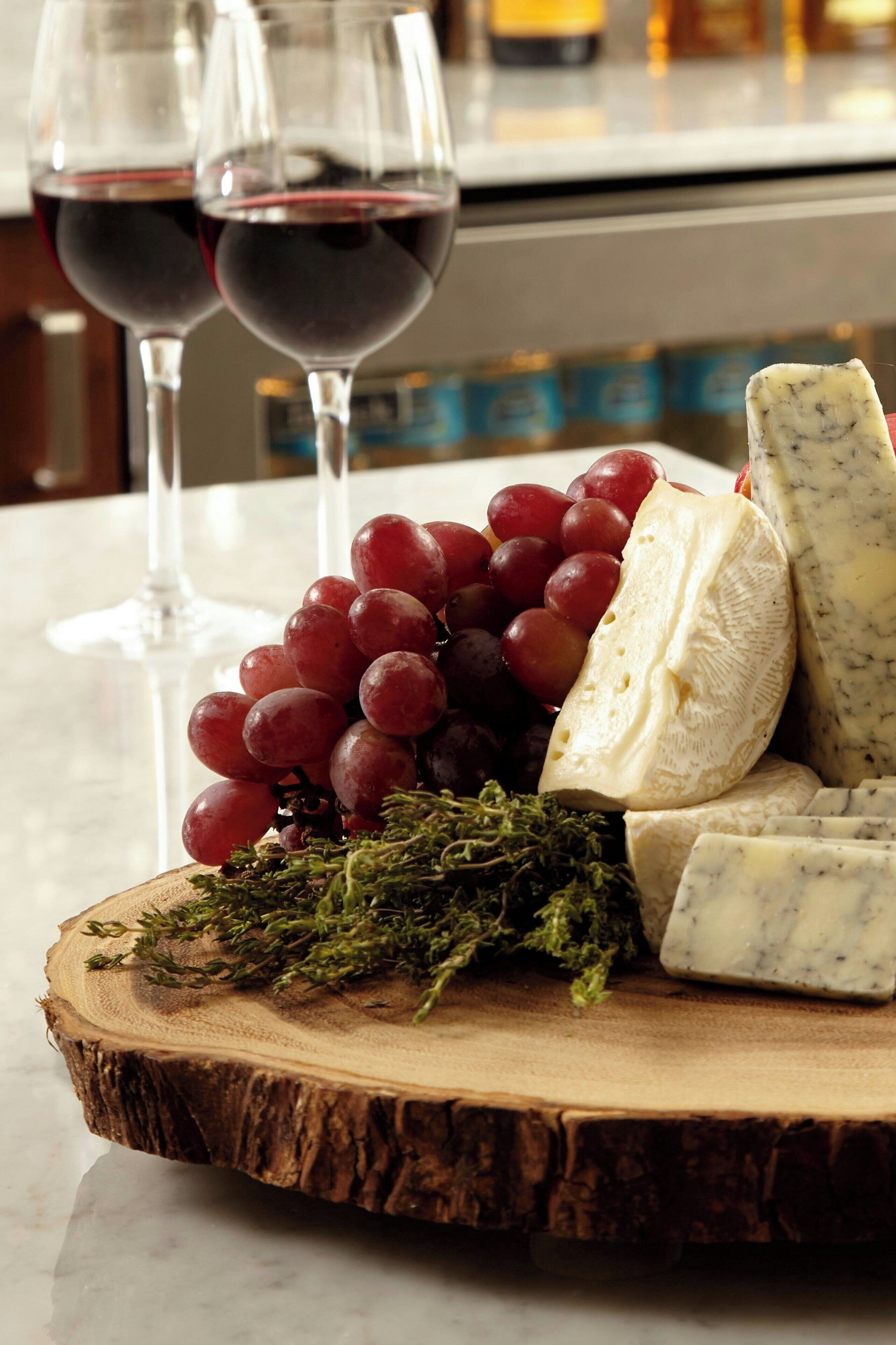 A wooden board with cheese, grapes, and herbs, and two glasses of red wine.