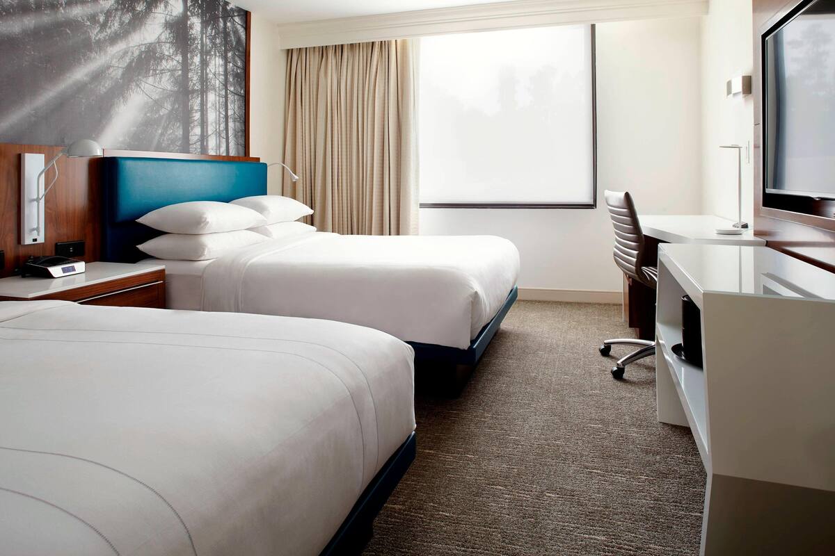A hotel room with two beds, a desk, a chair, a TV, and a large window with curtains.