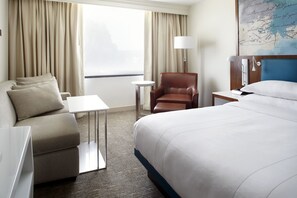 A hotel room with a bed, a sofa, a chair, a small table, a lamp, and a map on the wall.