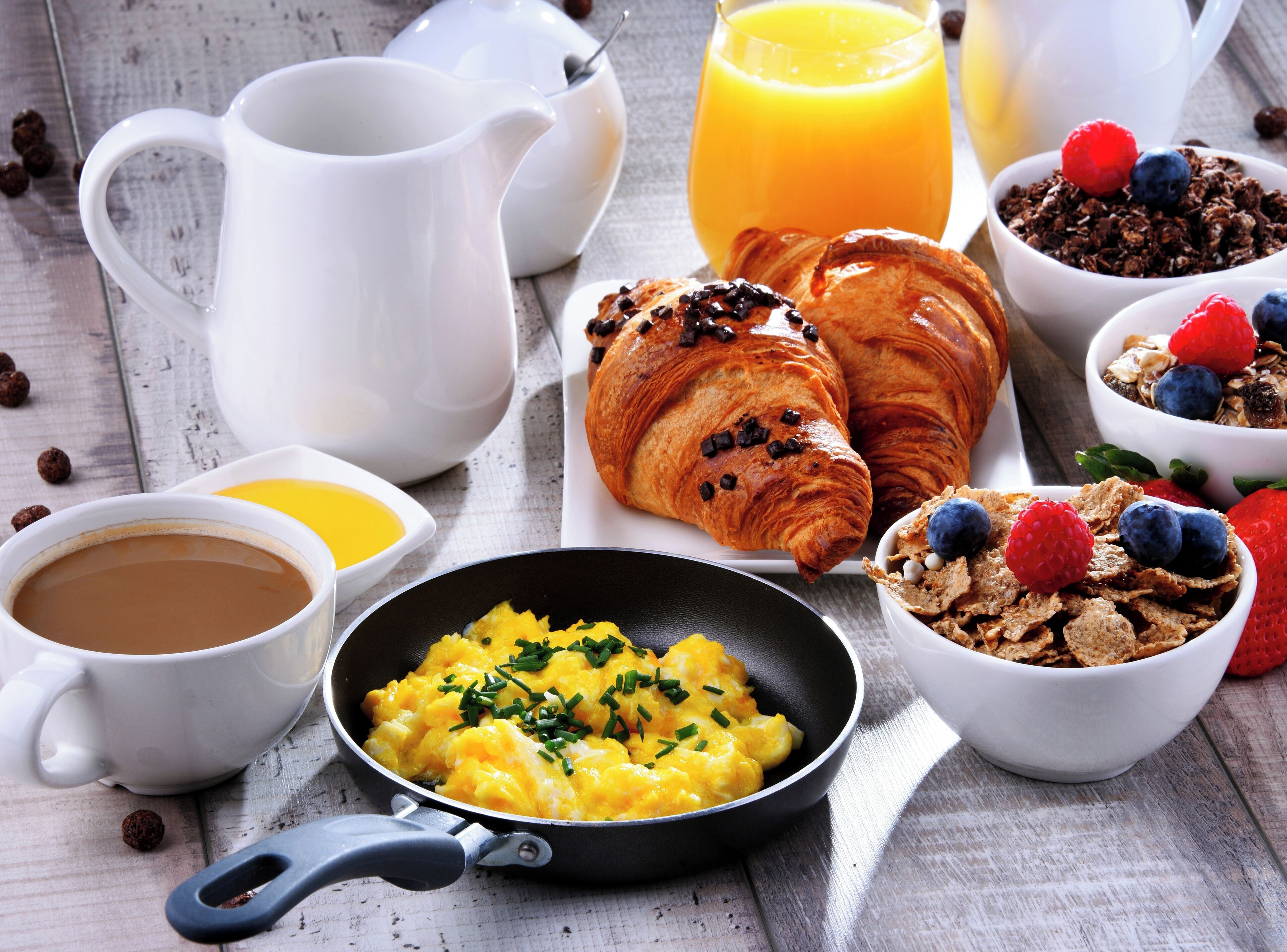 A breakfast setting with croissants, scrambled eggs, orange juice, coffee, and cereal with berries.