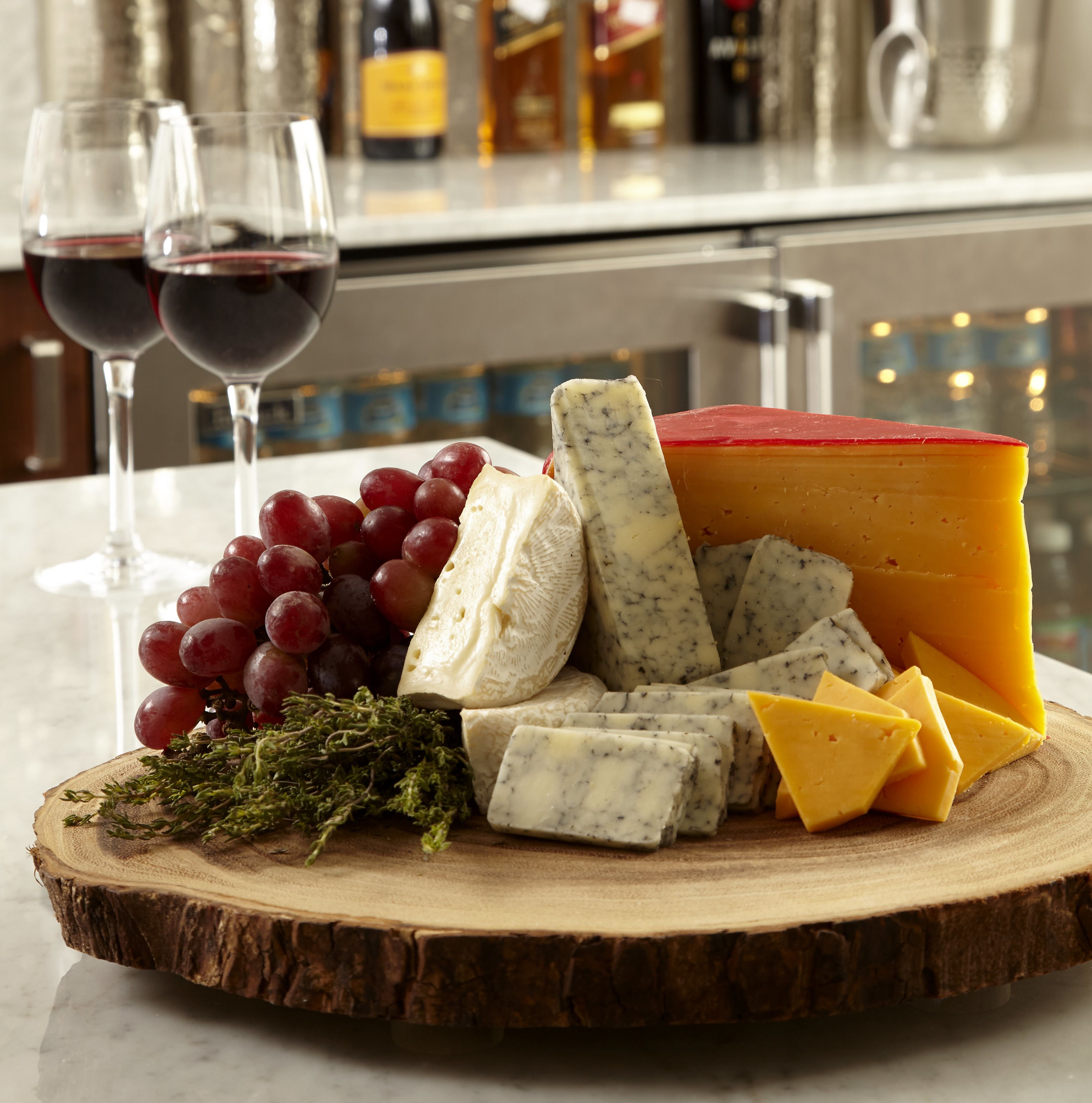 A wooden board with various cheeses, grapes, and a bottle of wine on a kitchen counter.