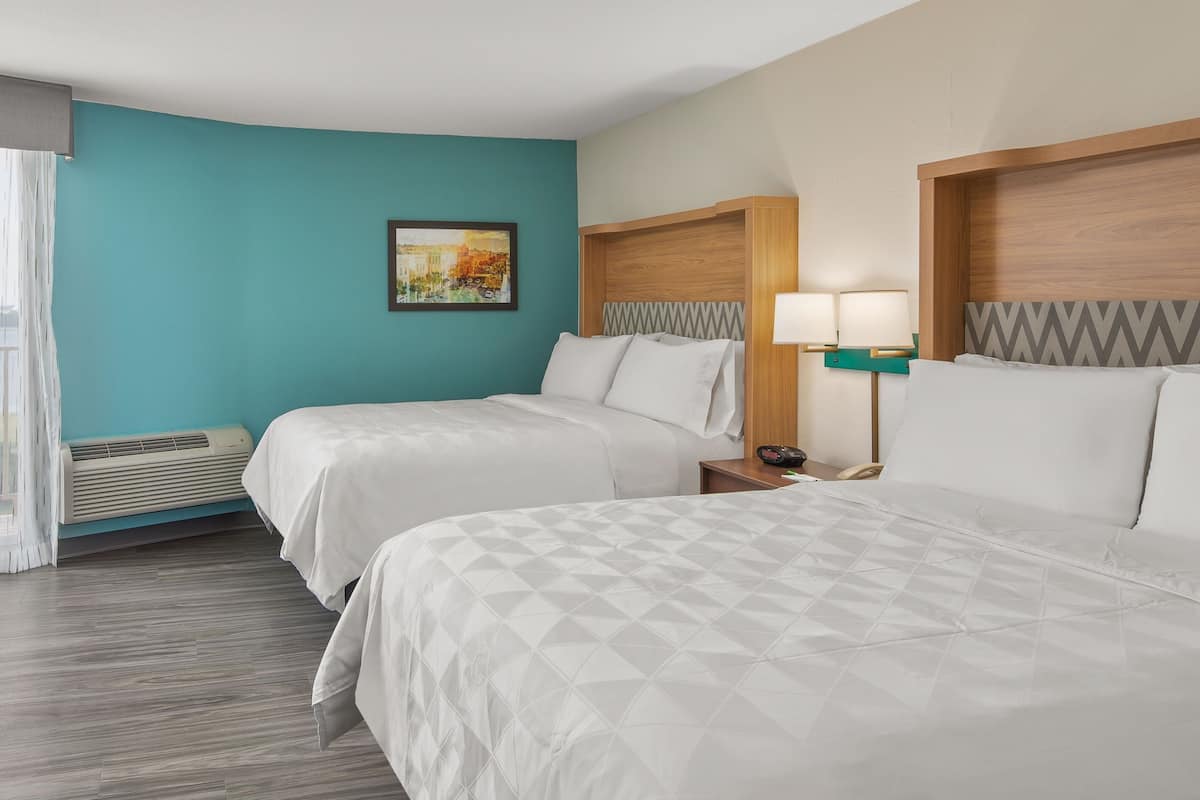 A hotel room with two beds, a painting on the wall, a nightstand with a lamp, and an air conditioning unit.
