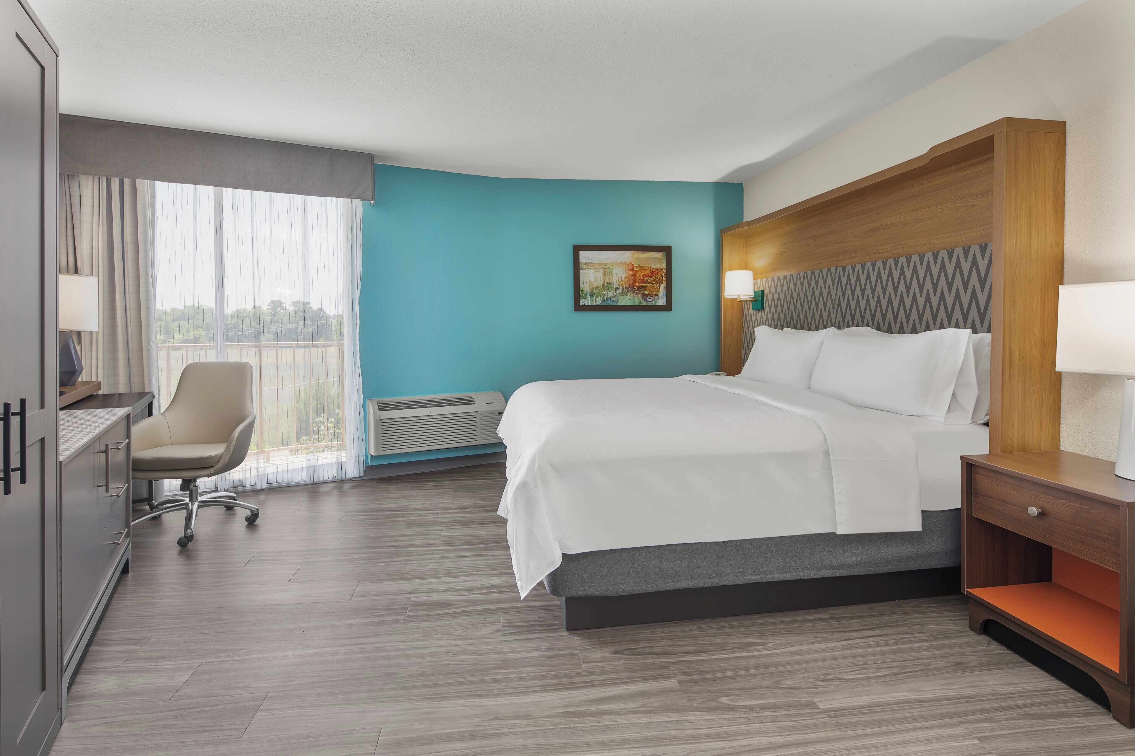 A hotel room with a bed, desk, chair, and a view of the outdoors.