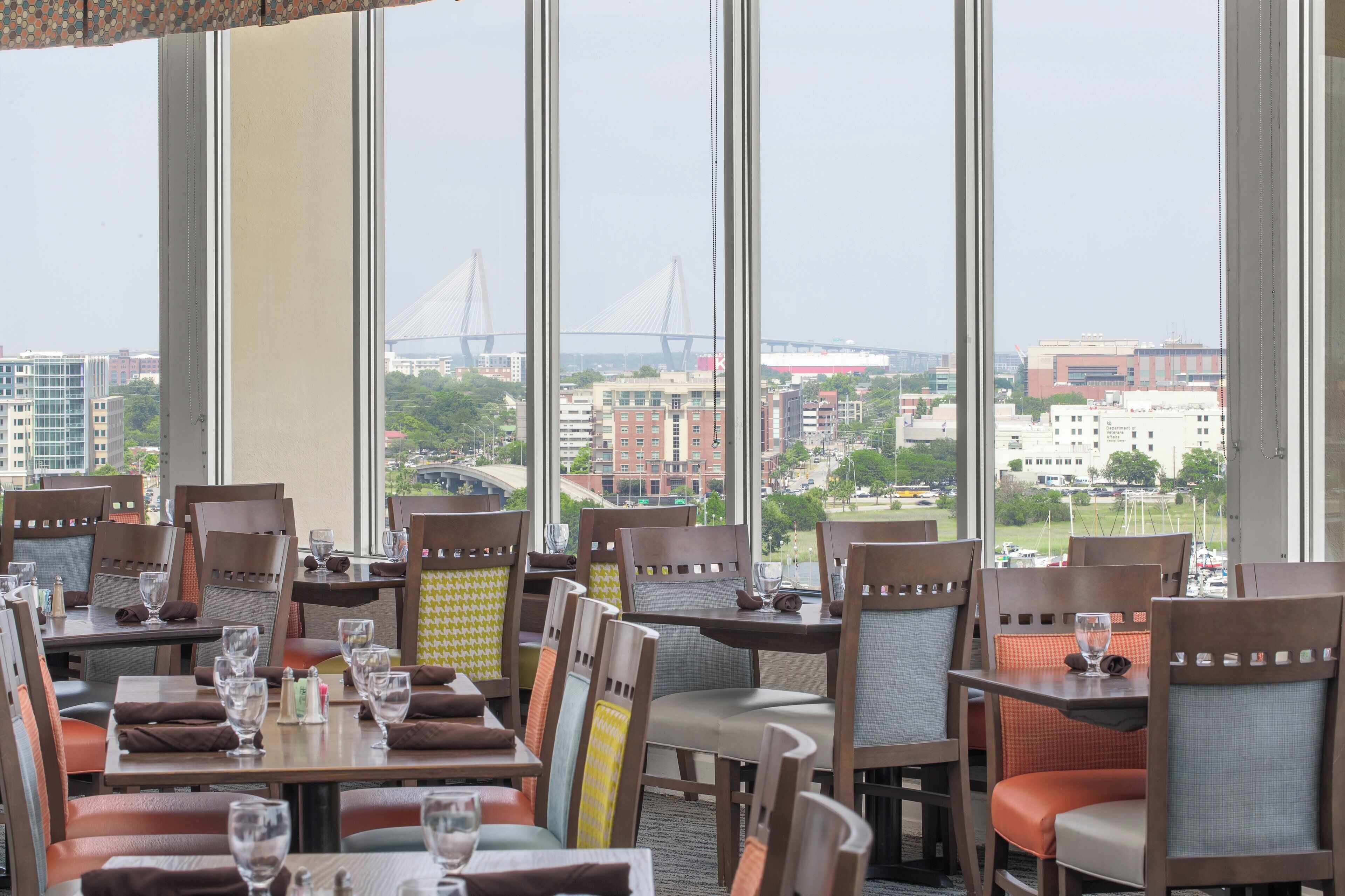 Un restaurant doté de nombreuses tables et chaises, de grandes fenêtres offrant une vue sur la ville et, au loin, sur un pont.