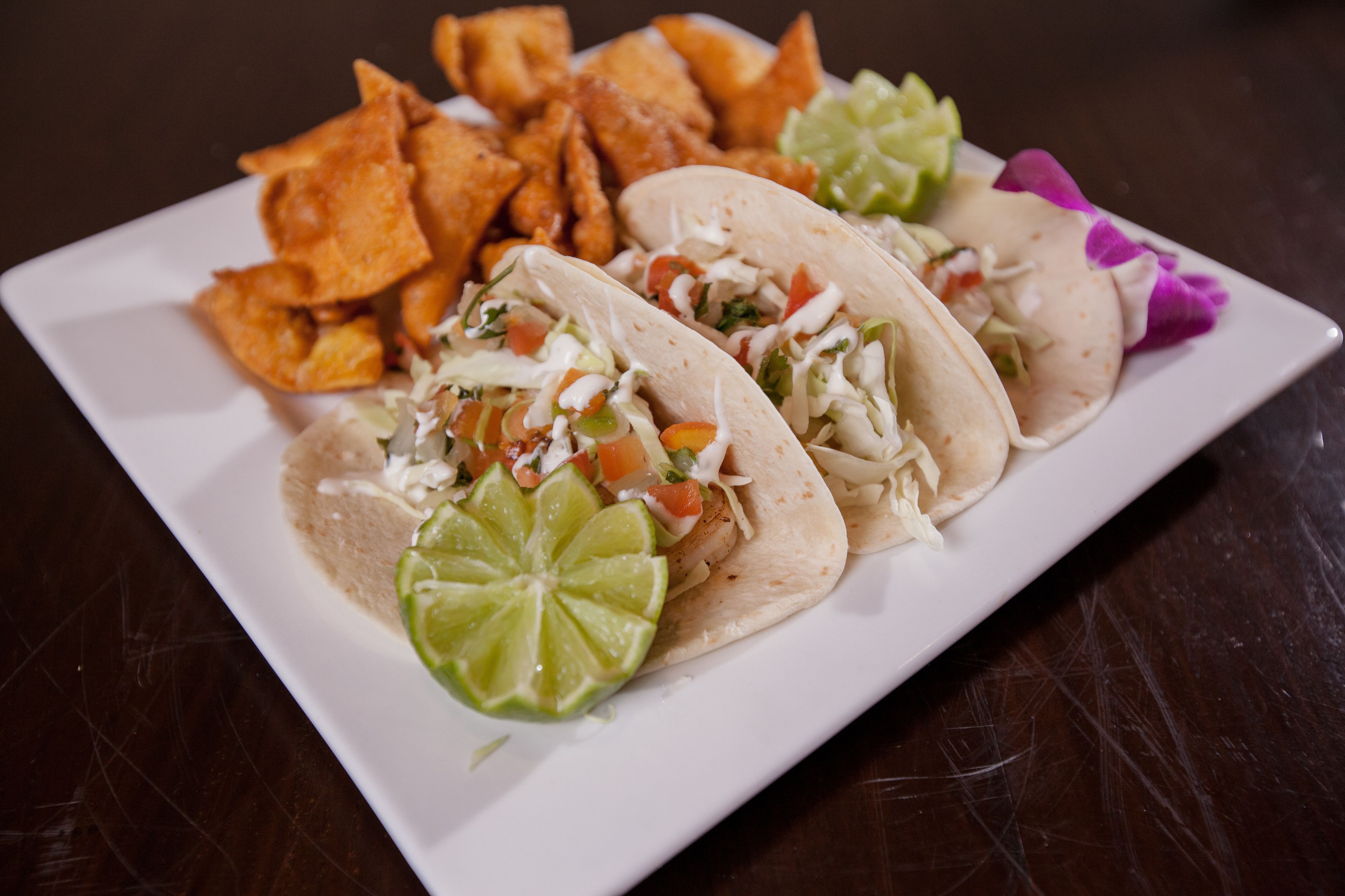 Three tacos with toppings and a side of tortilla chips on a white plate.