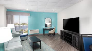 A hotel room with a large flat-screen TV, a sofa, a chair, a small table, and a window with a view of the city.