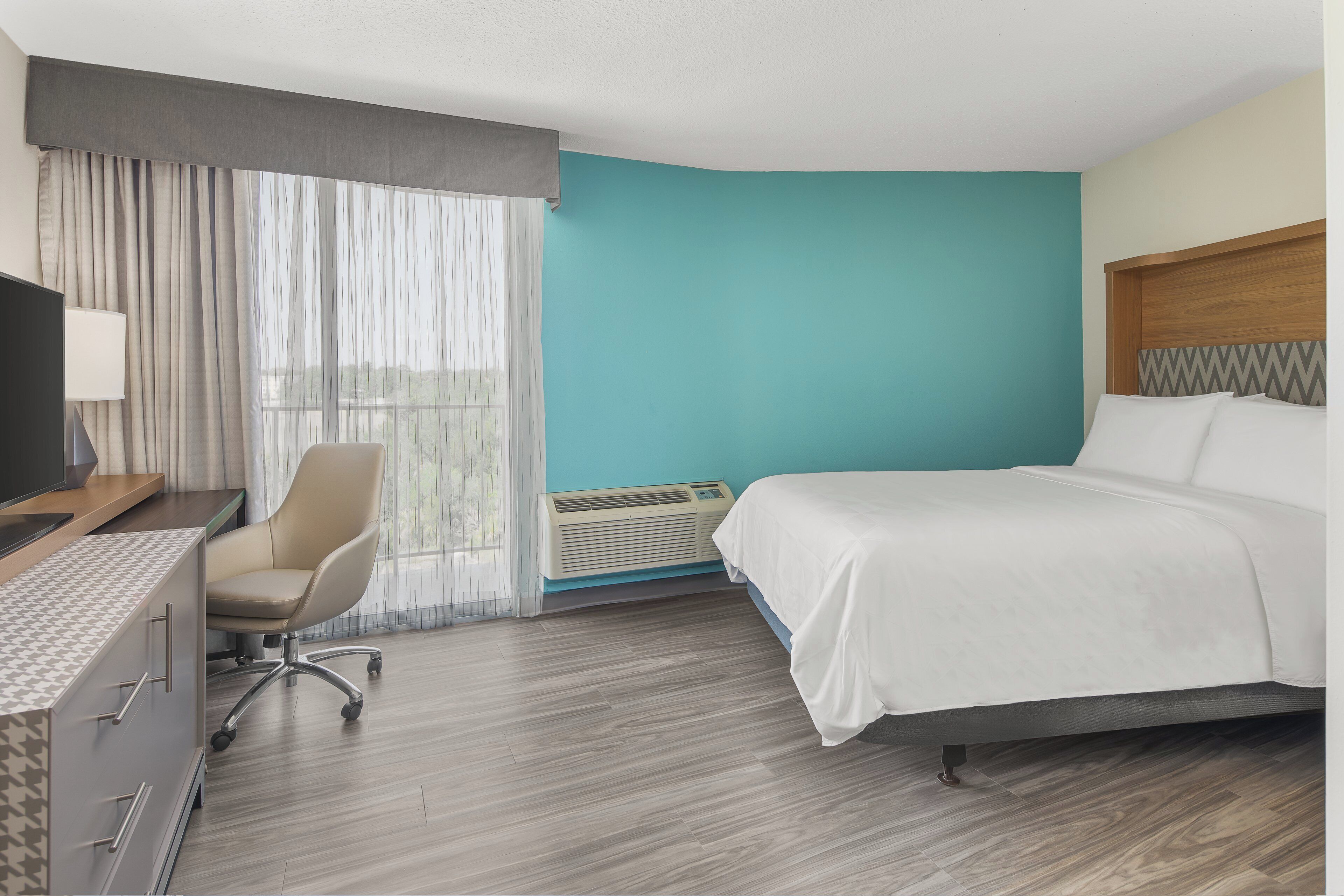 A hotel room with a bed, a desk, a chair, a TV, and a window with curtains.