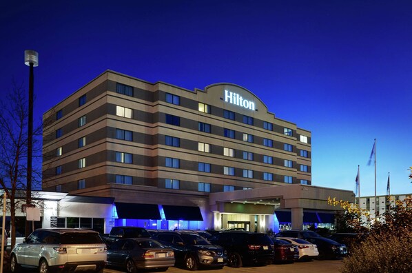 A multi-story hotel with a prominent 'Hilton' sign, a parking lot with several cars, and a clear sky.