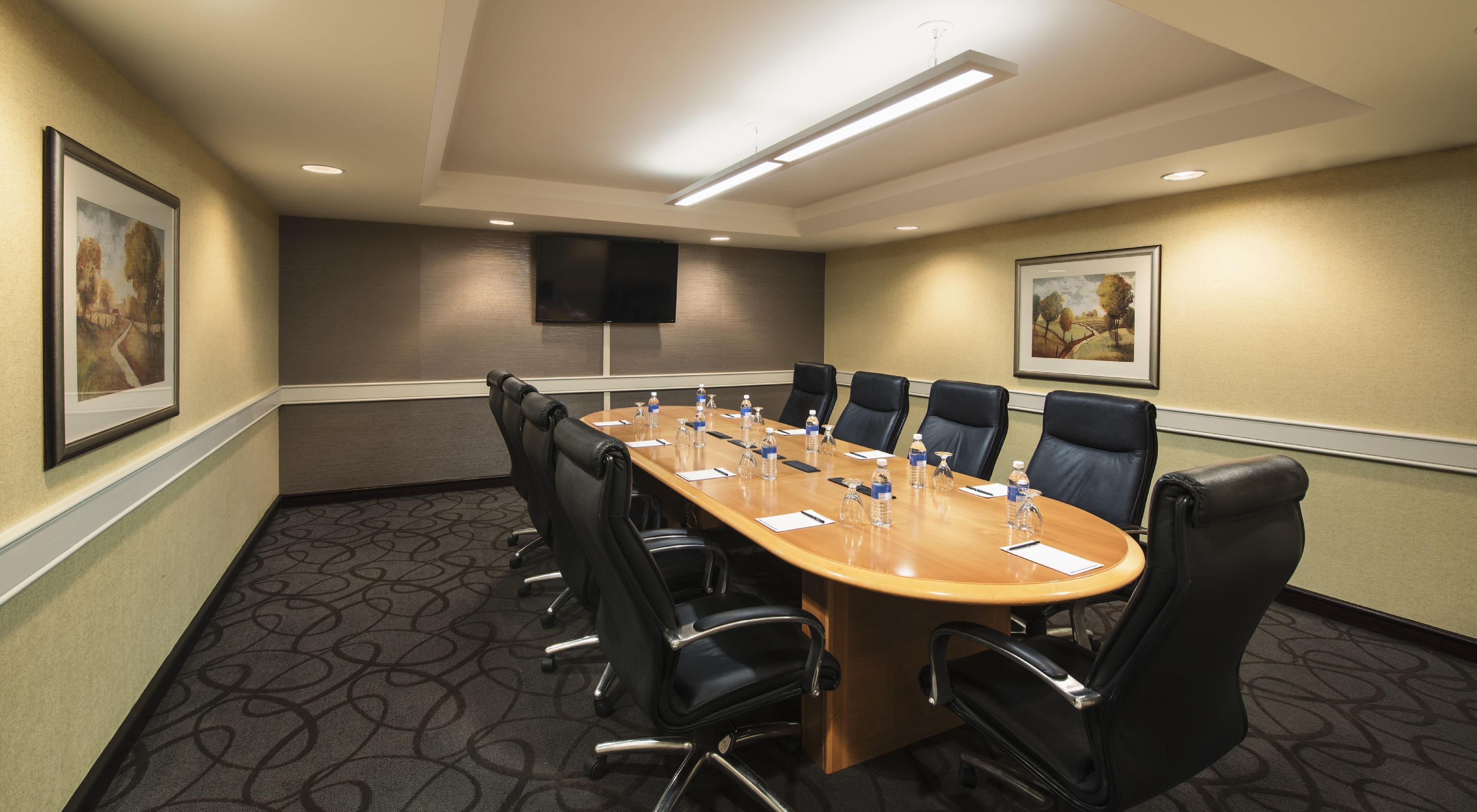 Une salle de conférence avec une longue table, des chaises noires, des bouteilles d’eau et un téléviseur à écran plat fixé au mur.