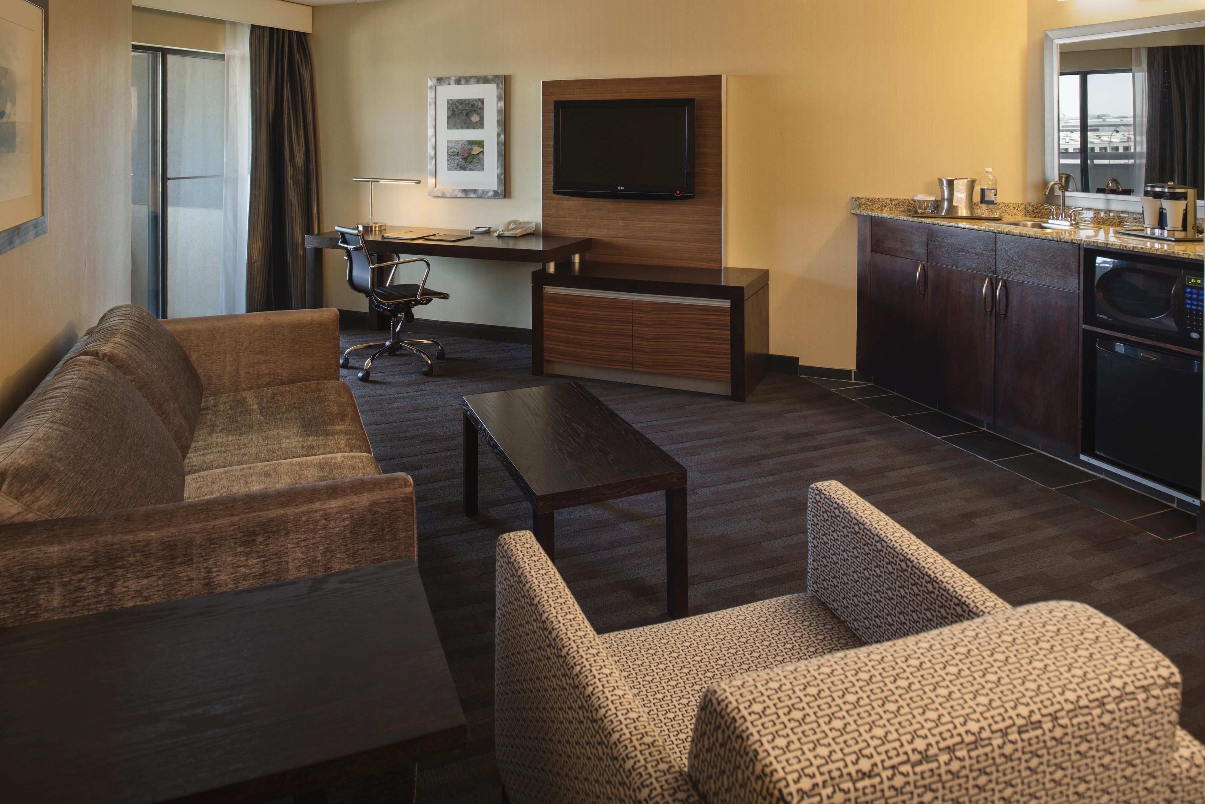 A hotel room with a sofa set, a desk with a chair, a flat-screen TV, a kitchenette, and a window with curtains.