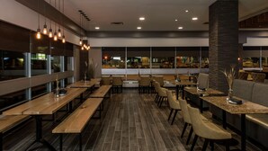 A modern restaurant with long wooden tables and benches, a stone pillar, and large windows offering a view of the street.