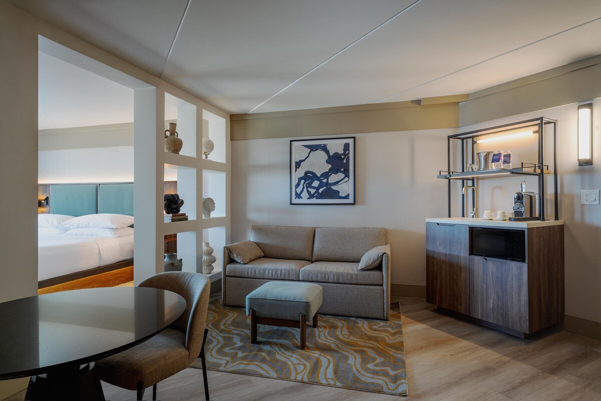 A compact hotel room with a bed, a sofa, a dining table, and a kitchenette.