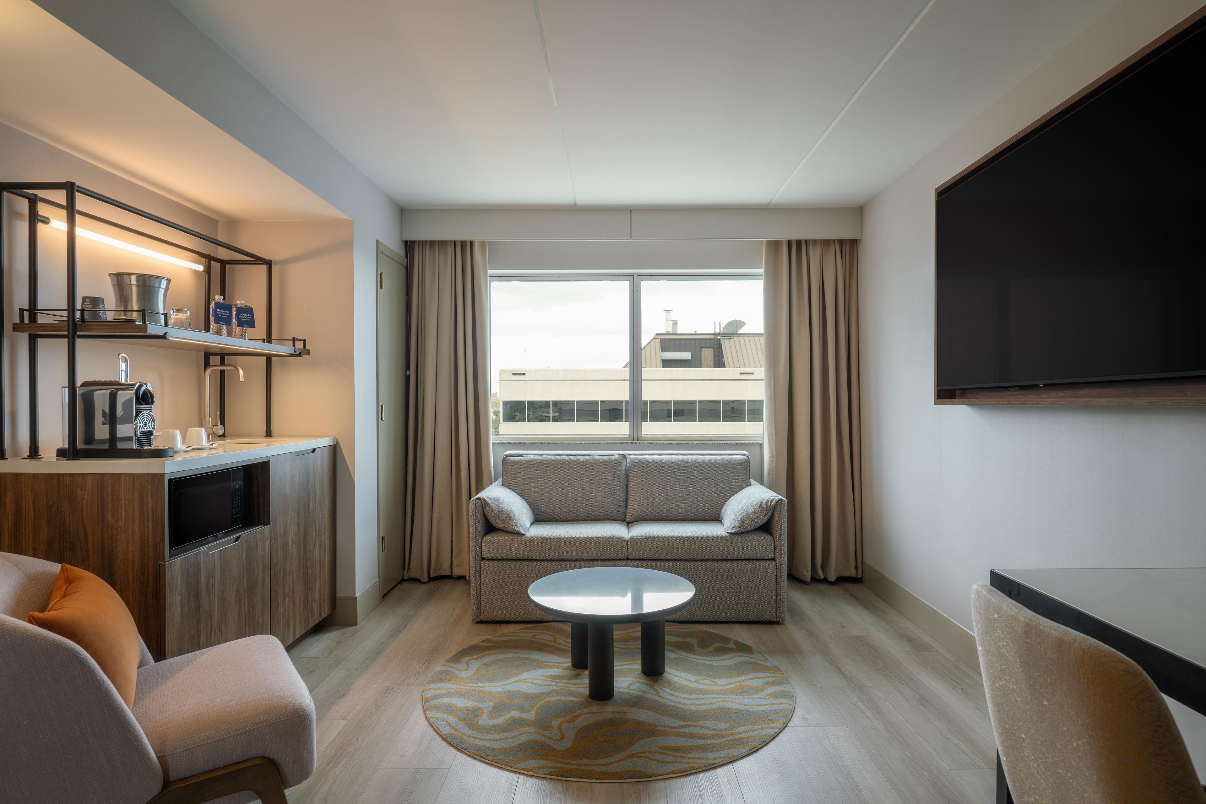 Una habitación de hotel moderna con sofá, una mesita redonda, una zona de cocina con cafetera y un televisor montado en la pared.
