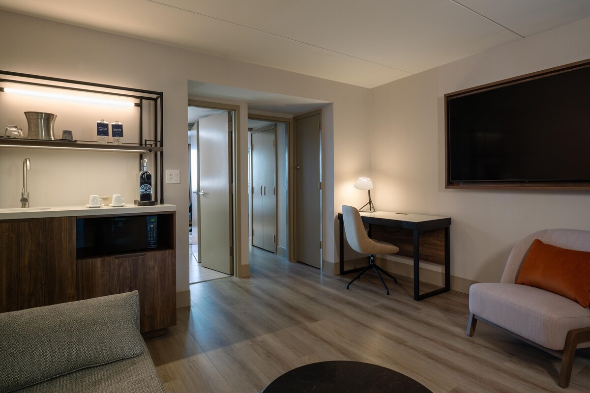 A hotel room with a kitchenette, a dining area with a table and chairs, a seating area with a sofa and armchair, and a large flat-screen TV mounted on the wall.