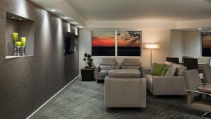 A modern hotel lobby with a seating area, a large window with a sunset view, and a flat-screen TV mounted on the wall.