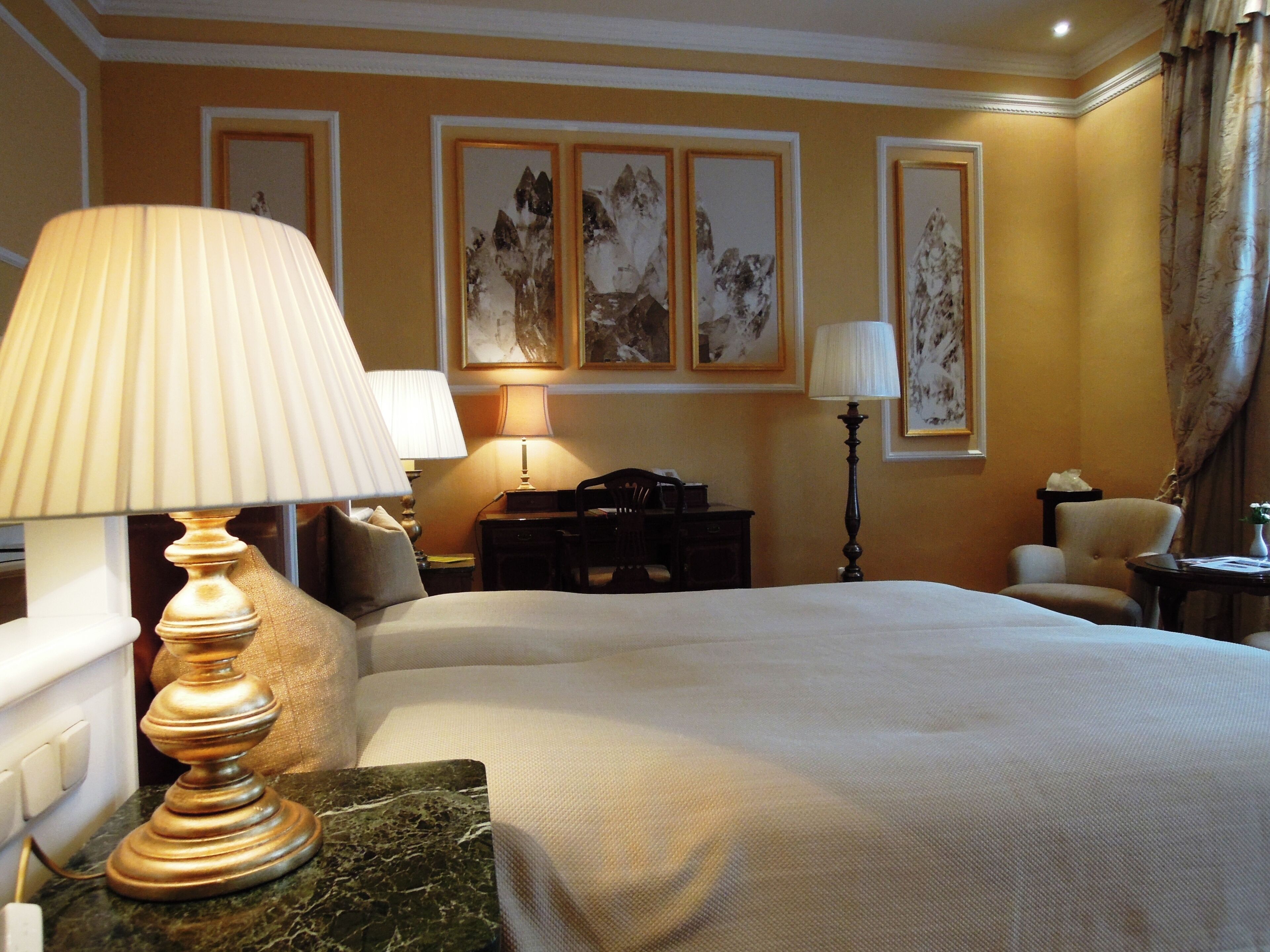 A hotel room with a bed, two bedside lamps, a desk, a chair, and artwork on the walls.
