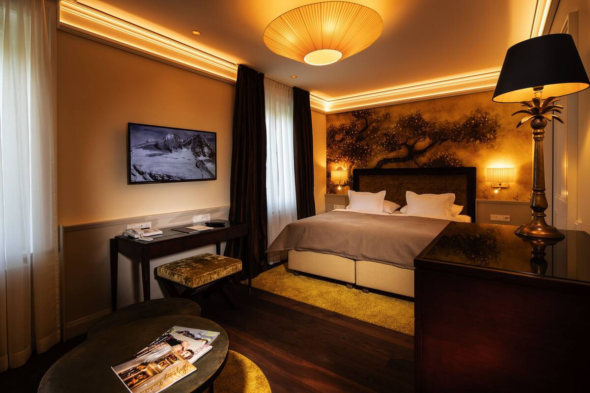 Ein Hotelzimmer mit Bett, Nachttischlampen, einem Schreibtisch mit Stuhl, einem runden Tisch mit einer Zeitschrift und einem Bild an der Wand.