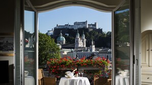 A dining area with a table set for two, overlooking a historic castle and lush greenery. - Bristol Hotel Salzburg (Salzburg)