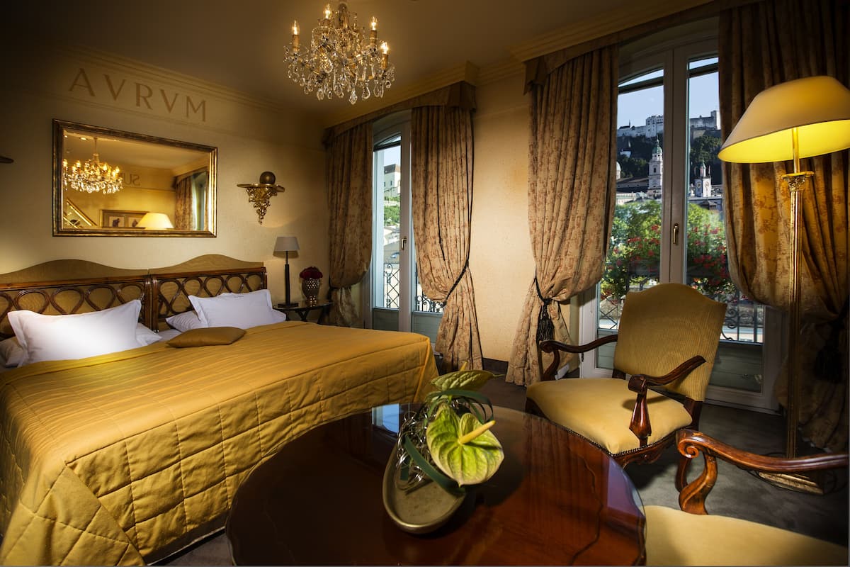 Ein Hotelzimmer mit Bett, Sessel, Tisch, einer Kronleuchter und Blick auf eine Stadtlandschaft durch das Fenster.