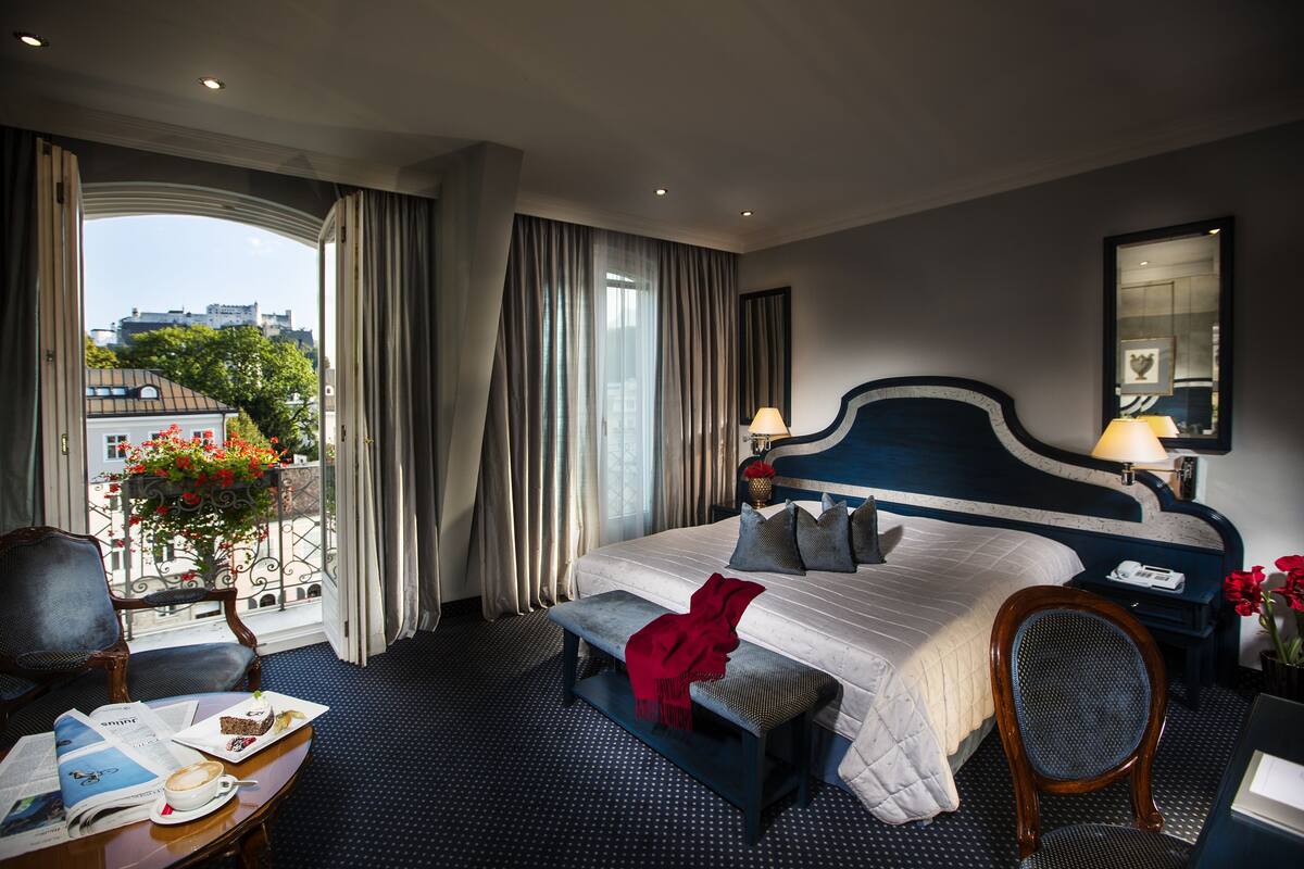 Ein Hotelzimmer mit einem großen Bett, einer Bank, einem Sessel, einem Tisch mit Zeitung und Tasse sowie Blick auf die Stadt durch ein Fenster.