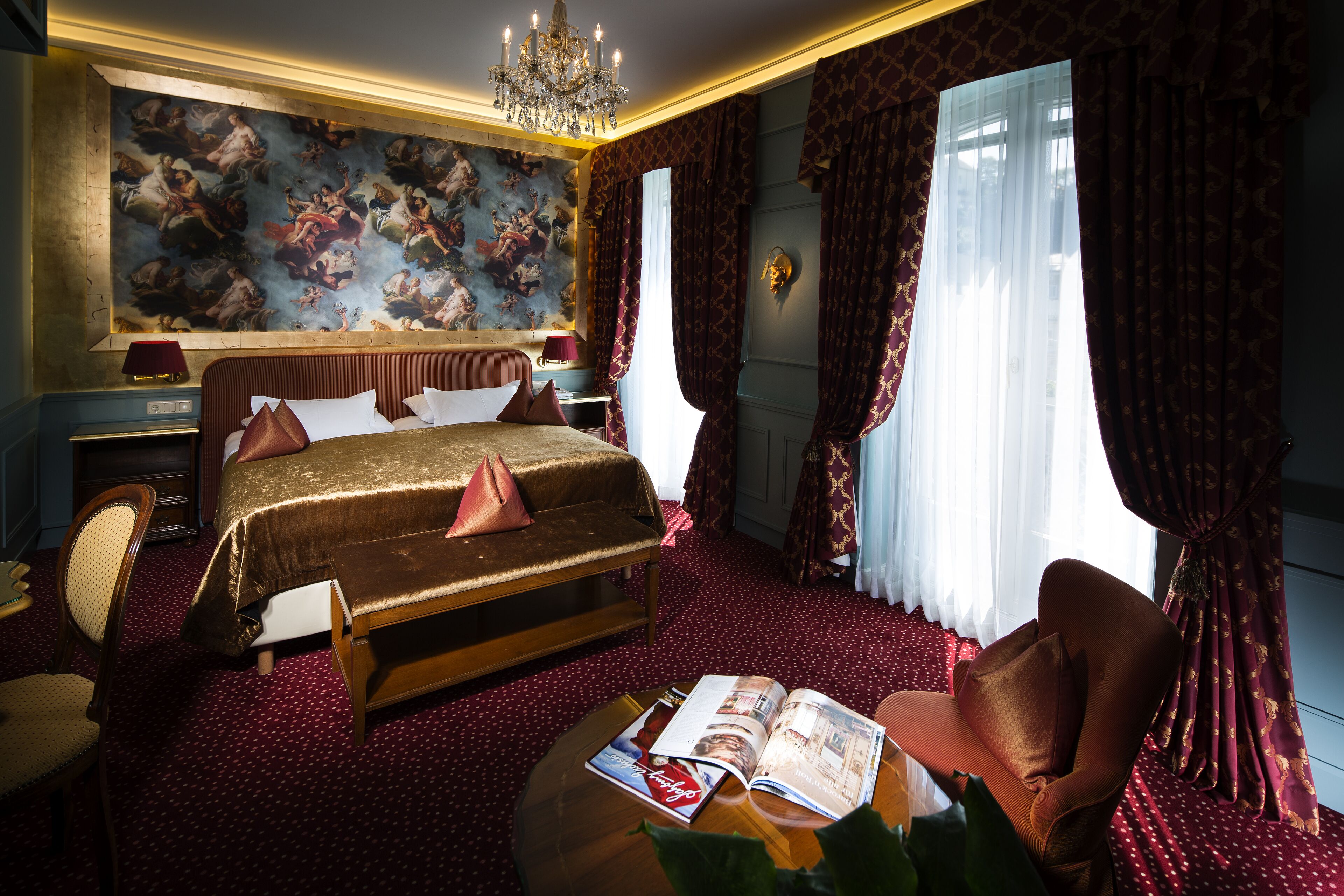 A luxurious bedroom with a large bed, a chandelier, a red carpet, and a large window with curtains.