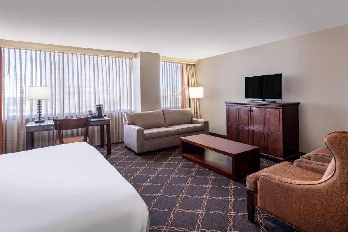 A hotel room with a bed, a sofa, a desk with a lamp, a television, and a cabinet.