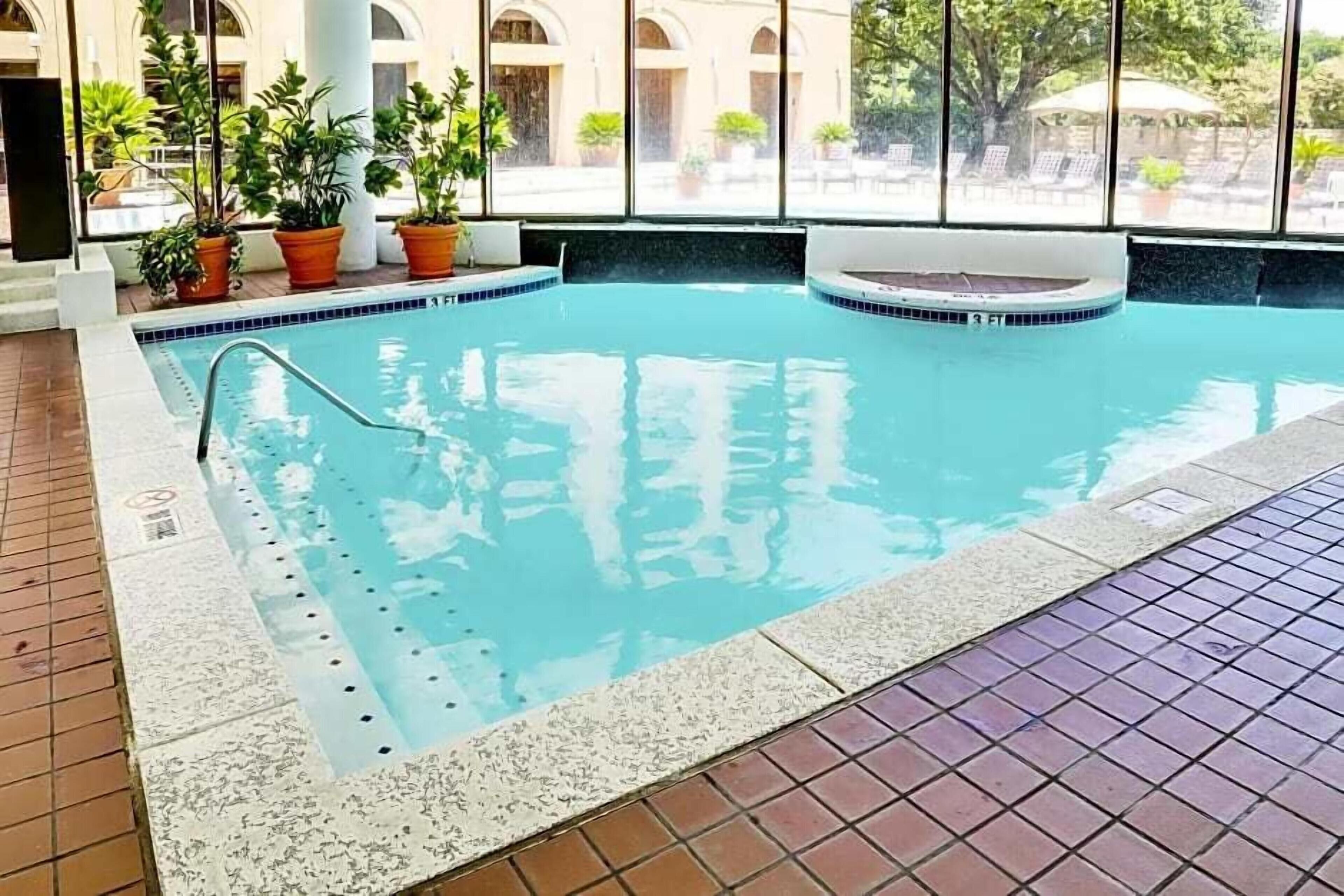An indoor swimming pool with clear water, a diving board, and potted plants around the perimeter.