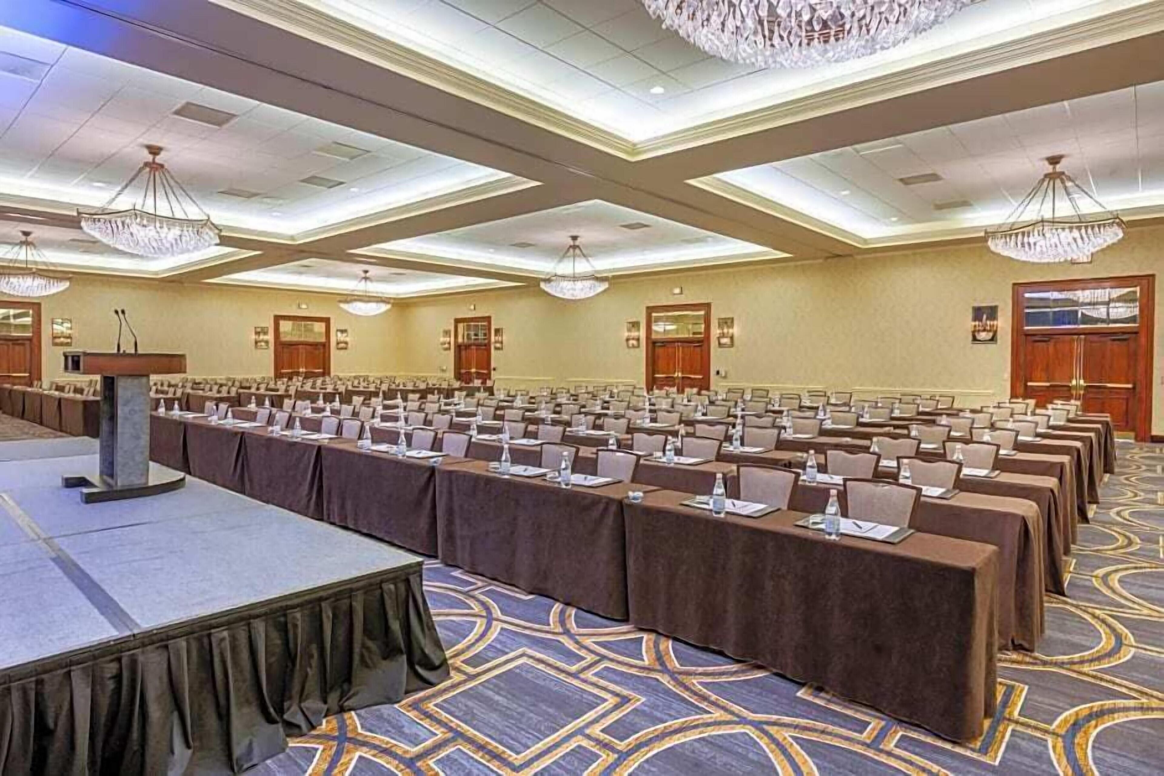 A conference room with rows of tables, a podium, and chandeliers.