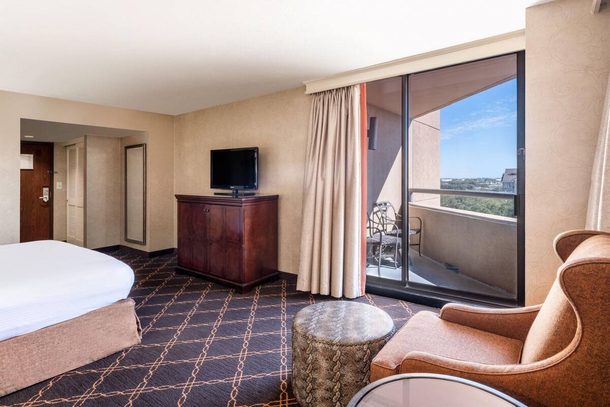 A hotel room with a bed, a TV on a wooden cabinet, a small round table, a chair, and a balcony with a view.