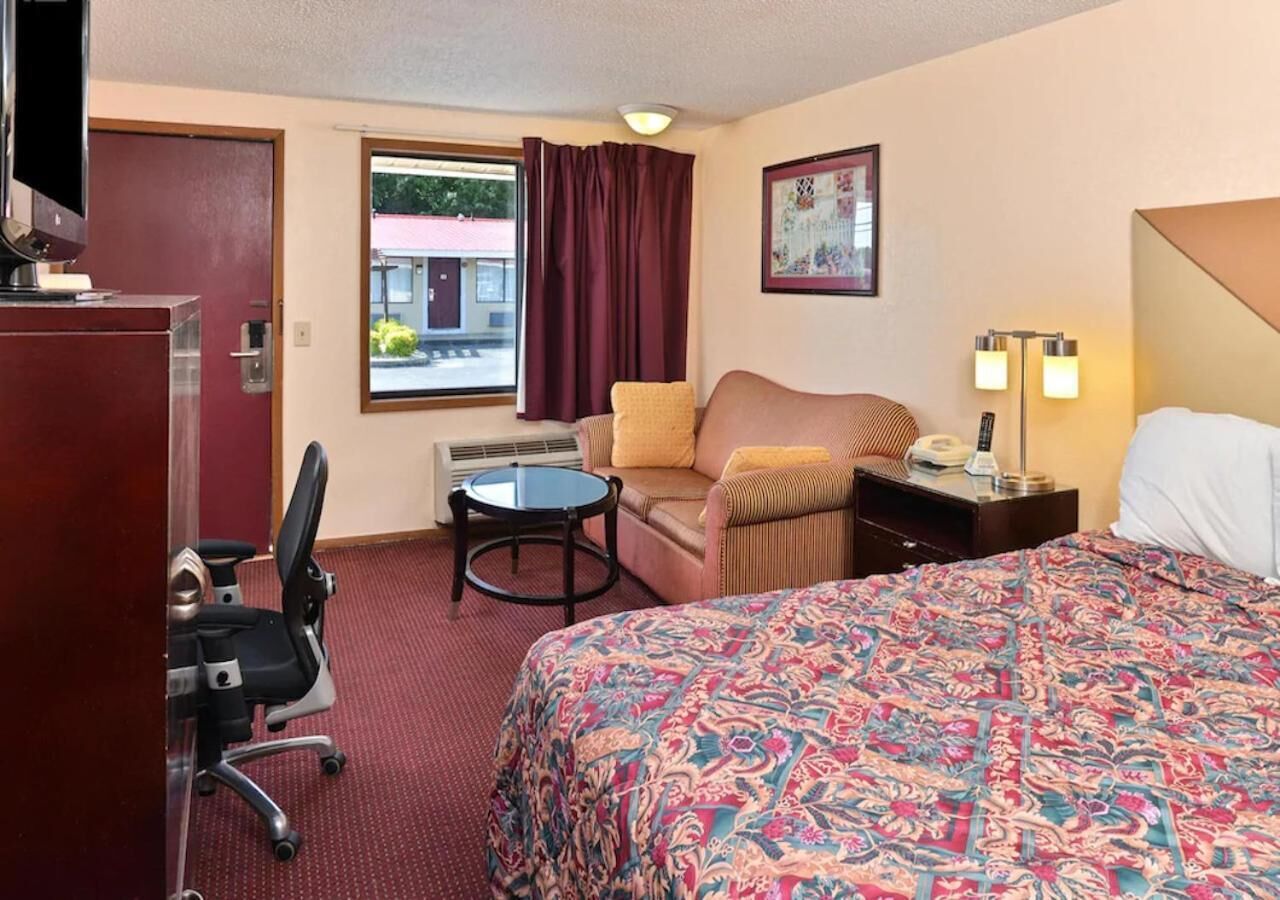 A hotel room with a bed, a desk, a chair, a small table, a TV, and a window with curtains.